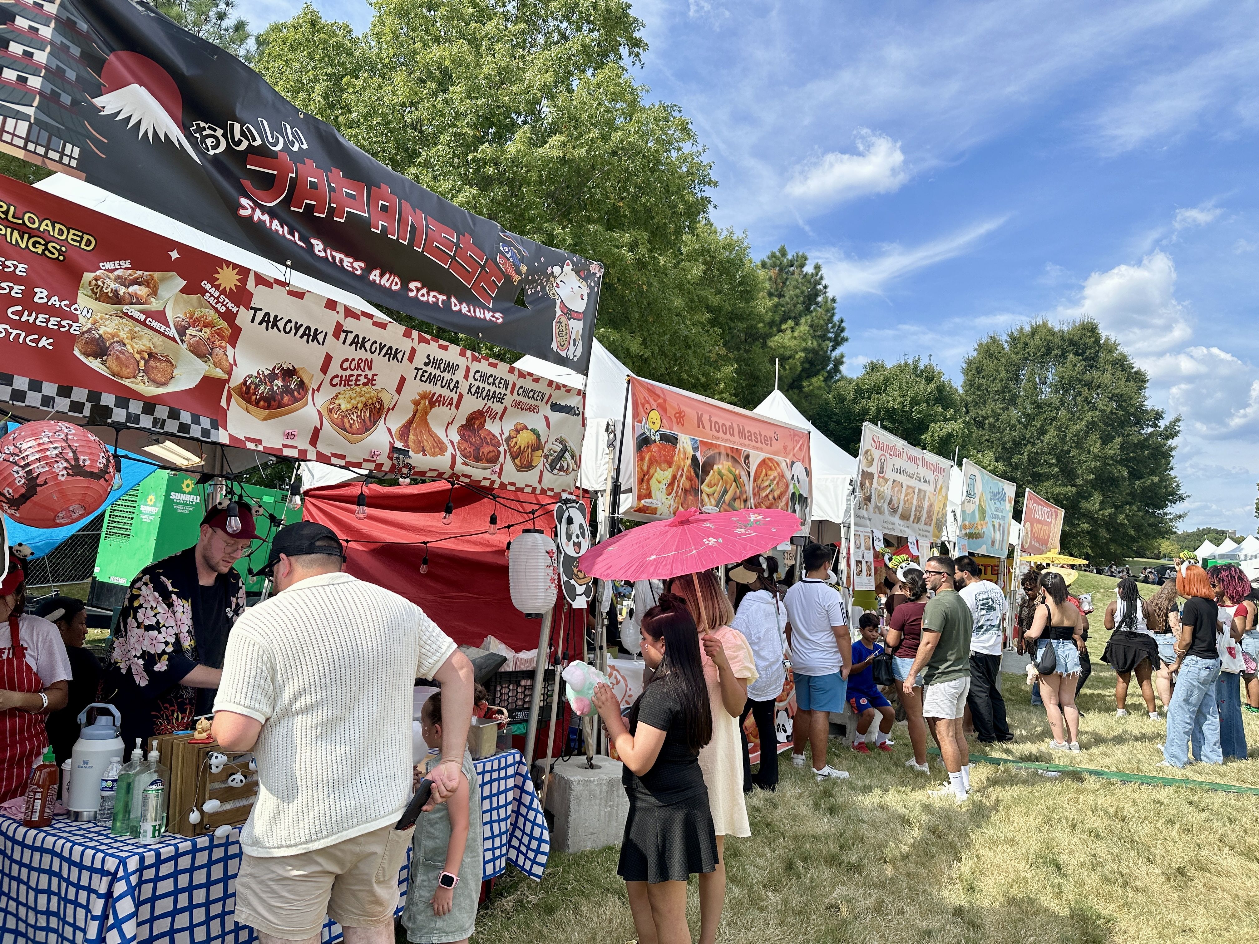 Outdoor food festival with multiple colorful food stalls including a Japanese stand selling takoyaki, shrimp tempura, and chicken karaage, people in casual summer attire waiting in line under a blue sky.