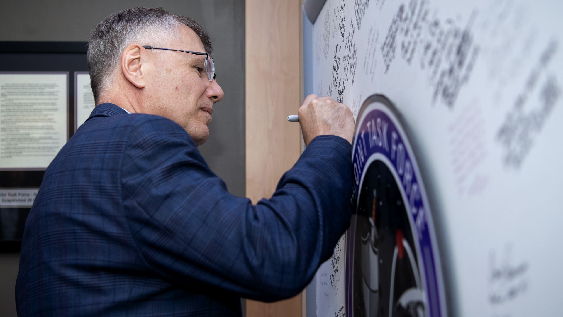 A man in a blue suit signs a decorative wall. Other signatures can be seen on the wall.
