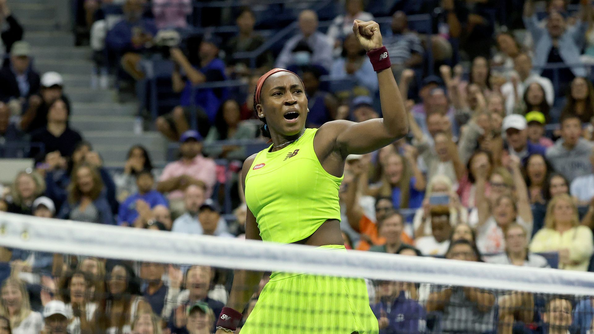 Coco Gauff wearing bright green with spectators in the background. She stands in front of the tennis net with a fist in the air 