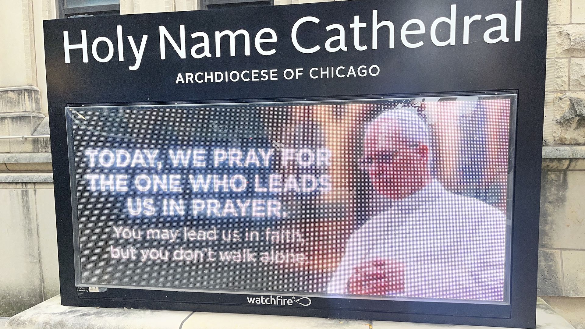 Holy Name Cathedral is written in large white letters on a black panel, with Archdiocese of Chicago beneath. A digital display shows praying text about the one who leads us, with a Watchfire logo.