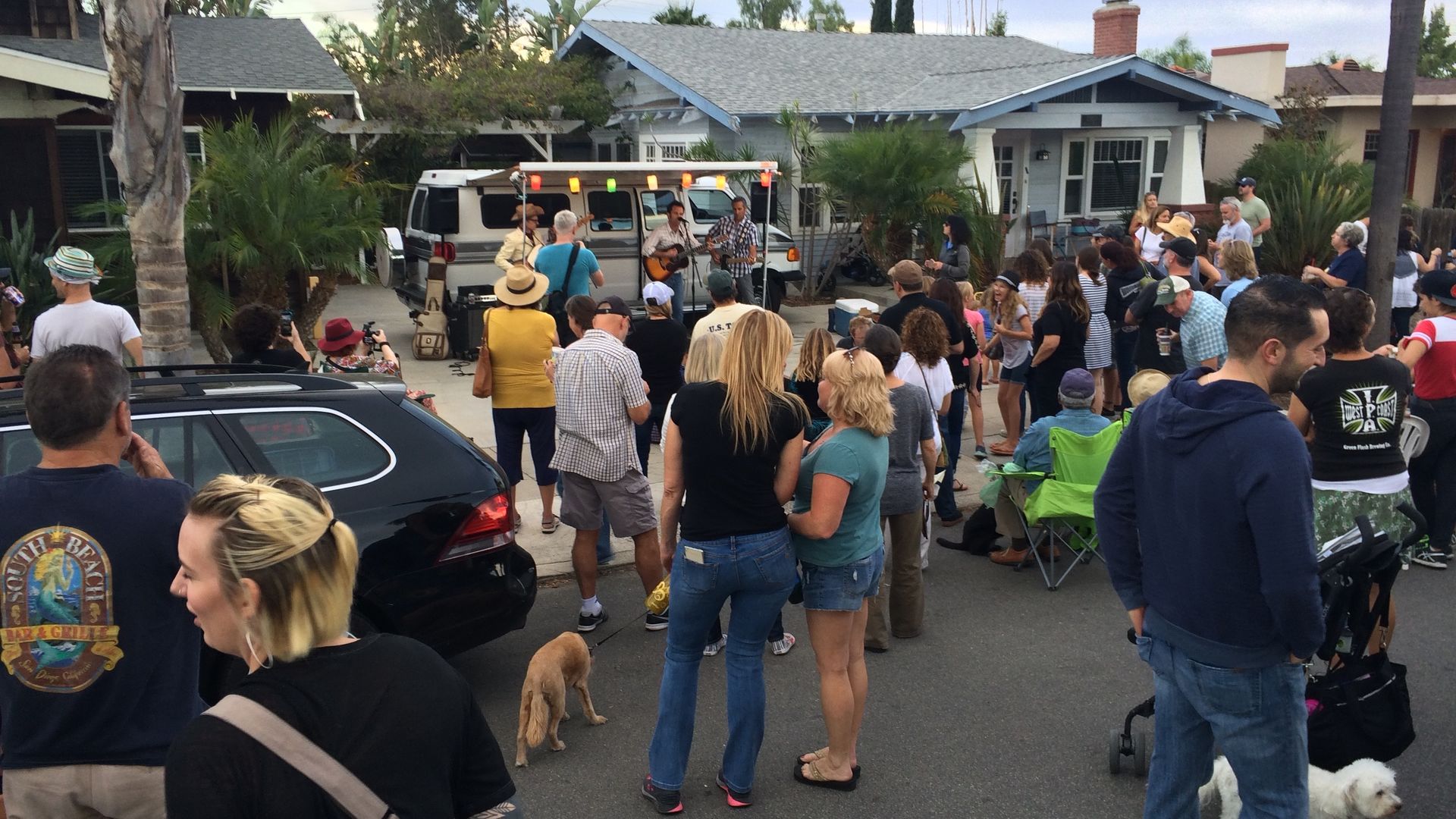 A small crowd gathers in the street while a band plays under an awning drawn from a camping van parked in a driveway