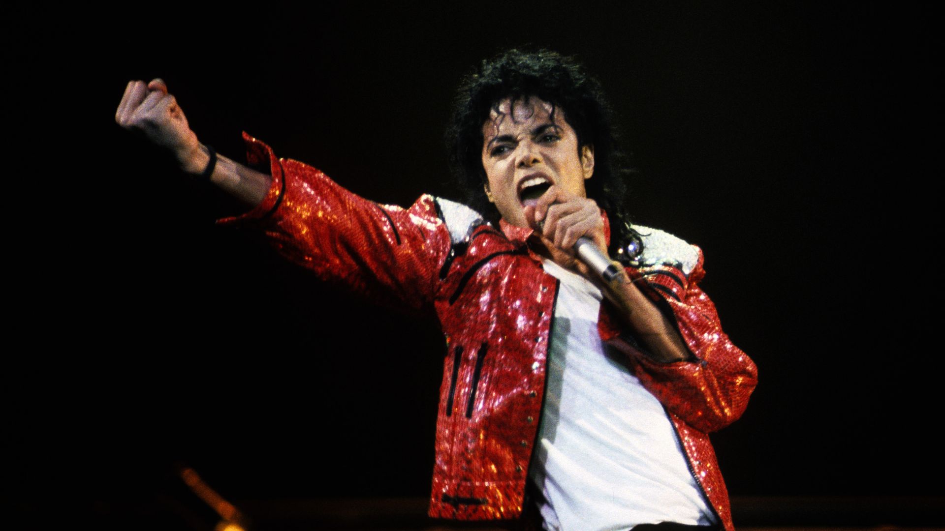 A male pop star performs on stage in a bright red sequined jacket, white shirt, and black pants, singing into a microphone with his arm raised against a dark background.