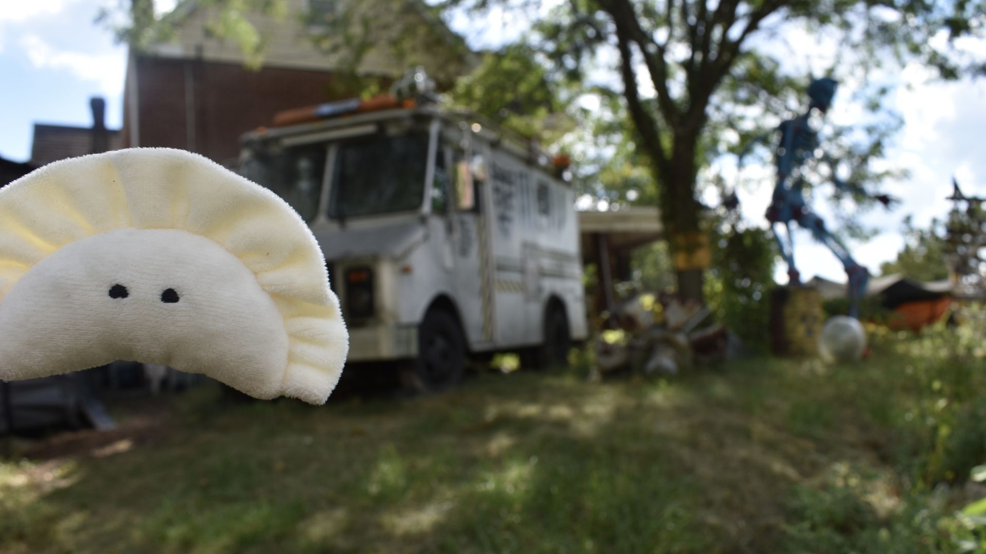 Plush toy resembling a smiling white dumpling held close to the camera, with an old white truck and a blue metal skeleton sculpture in a grassy yard blurred in the background.