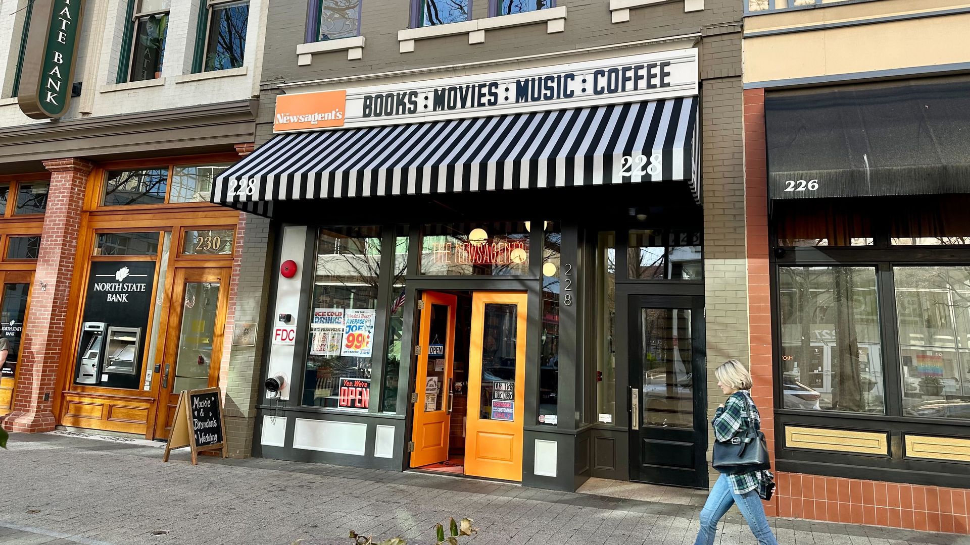 Street view of a bookstore with a black and white striped awning and orange doors at 228. Sign reads Newsagent's books, movies, music, coffee. Woman in jeans and plaid shirt walks by.