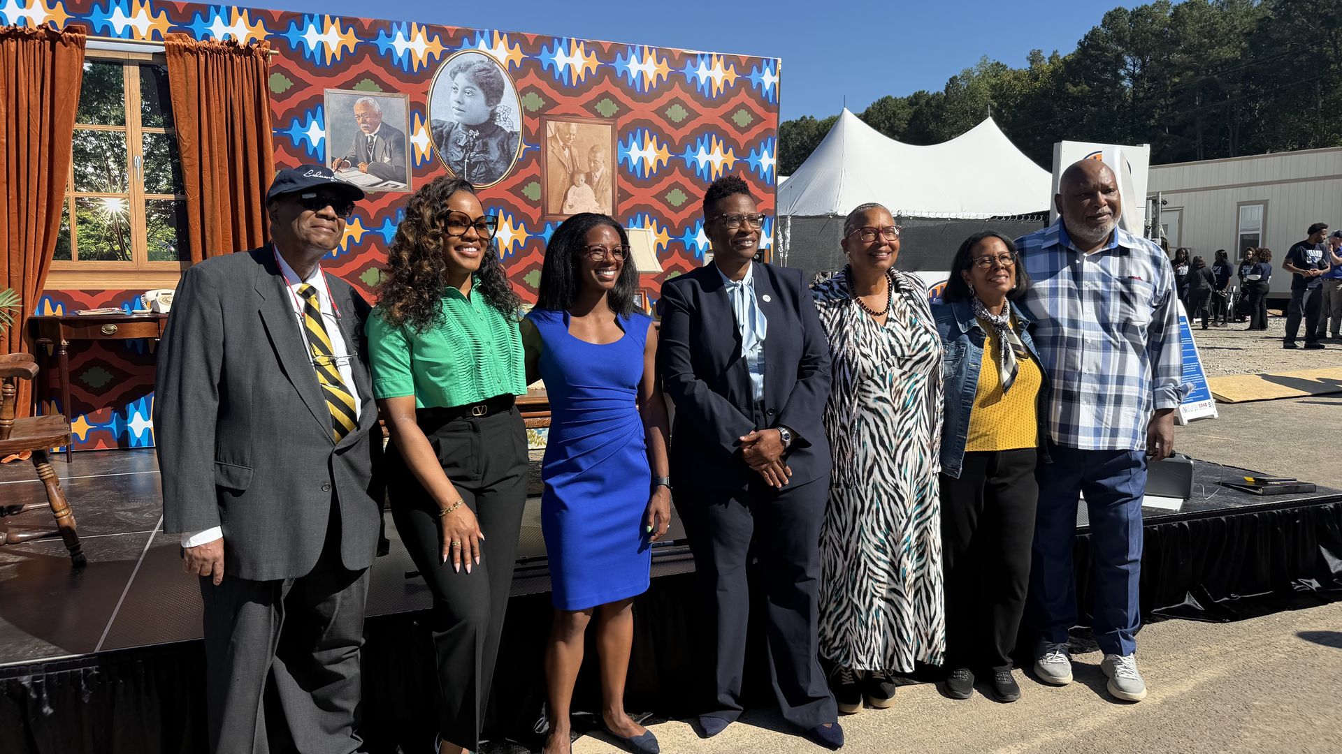 Seven people pose outdoors in front of a vibrant wall mural with framed portraits; a stage with brown curtains on the left and a white event tent in the background; they smile for the photo.