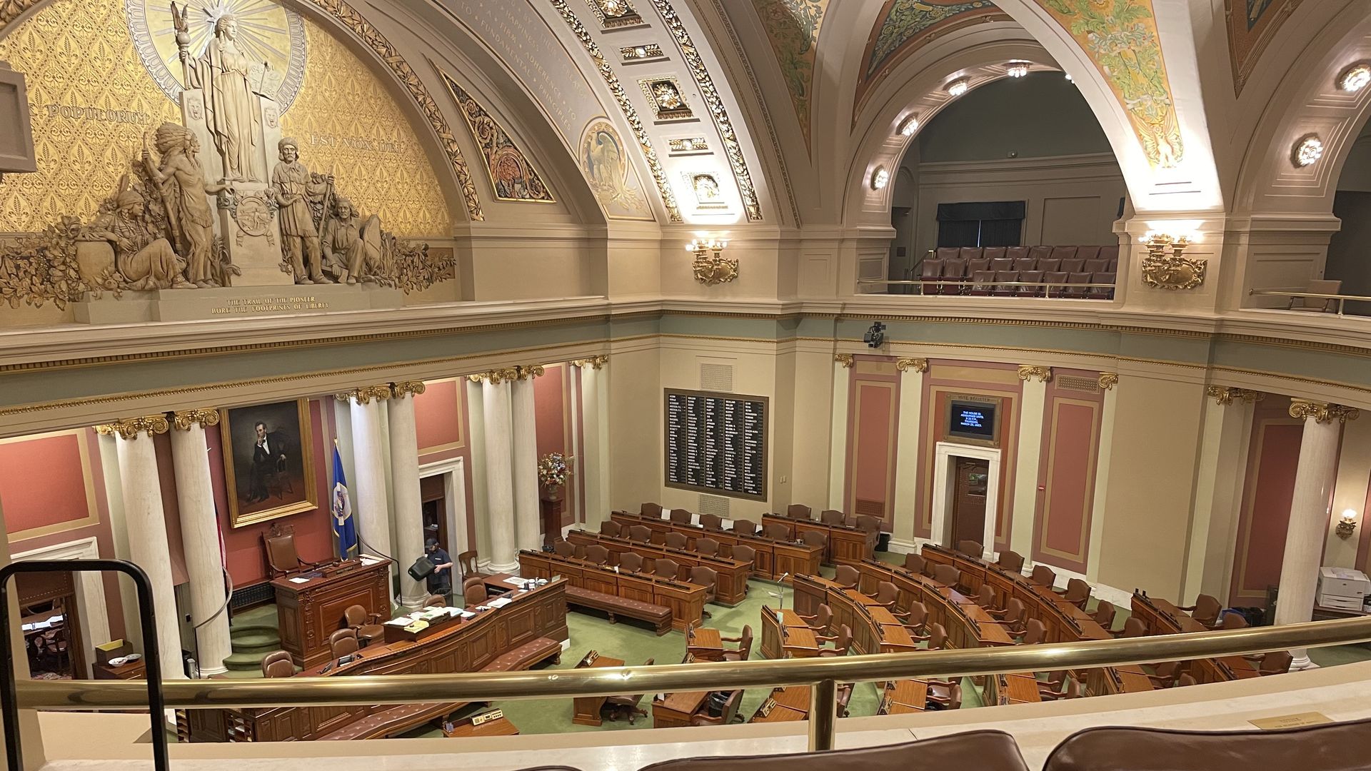 Interior view of a legislative chamber with rows of wooden desks and chairs, ornate gold and red walls, a bas-relief sculpture, and a balcony with leather seats in the foreground.