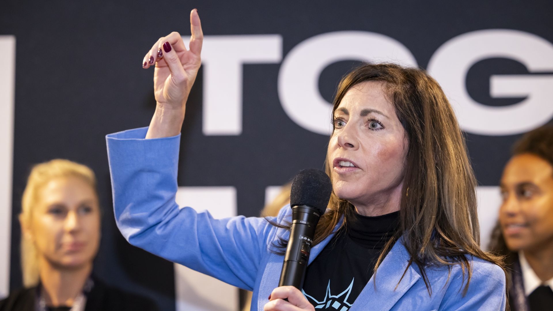 New Jersey First Lady Tammy Murphy, wearing a black t-shirt and powder blue blazer, and holding a microphone, surrounded by supporters.