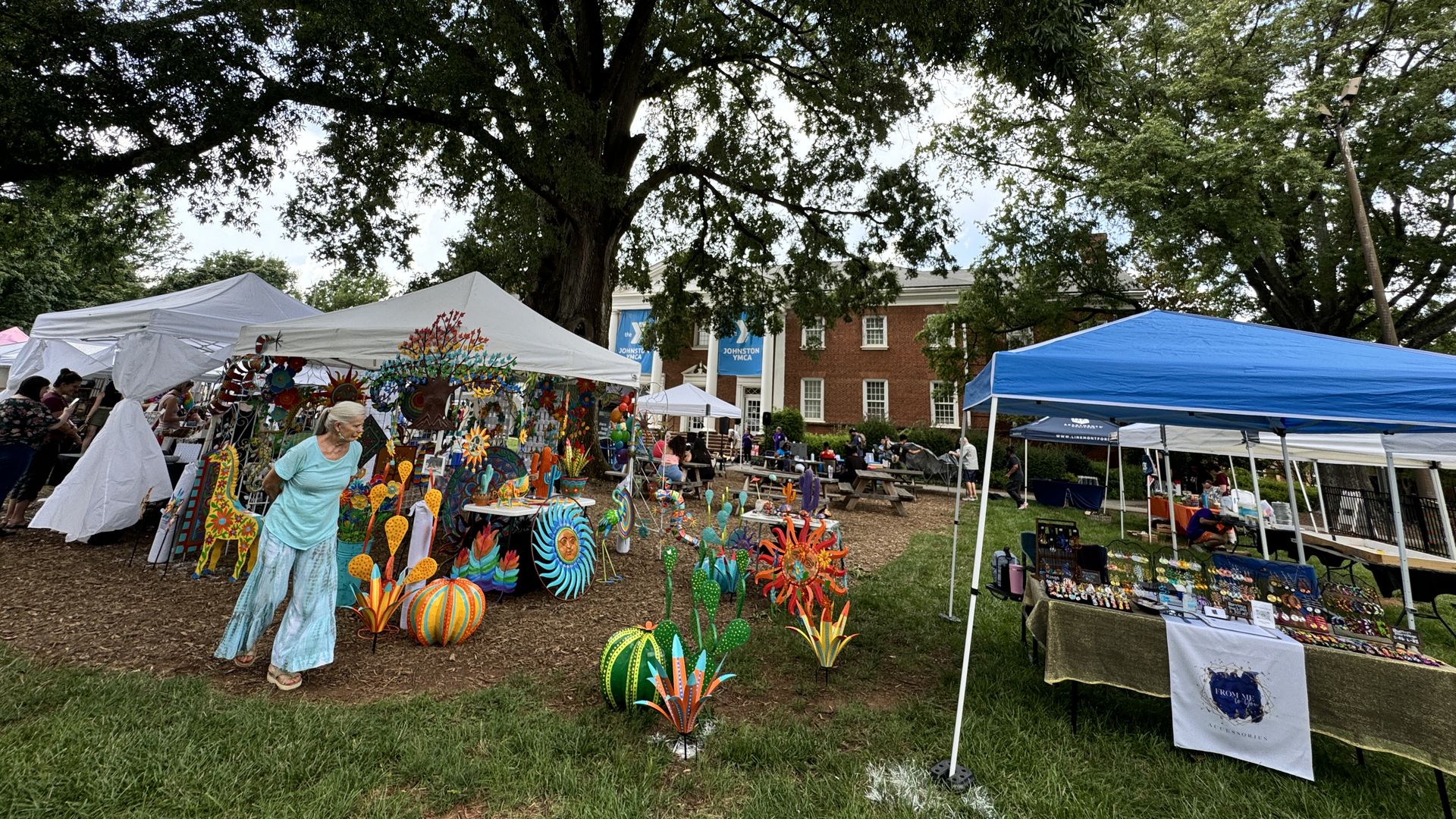 Outdoor craft fair with colorful metal garden decorations under large trees. White tents display various art, and people browse near a red brick building with "Johnston YMCA" banners.