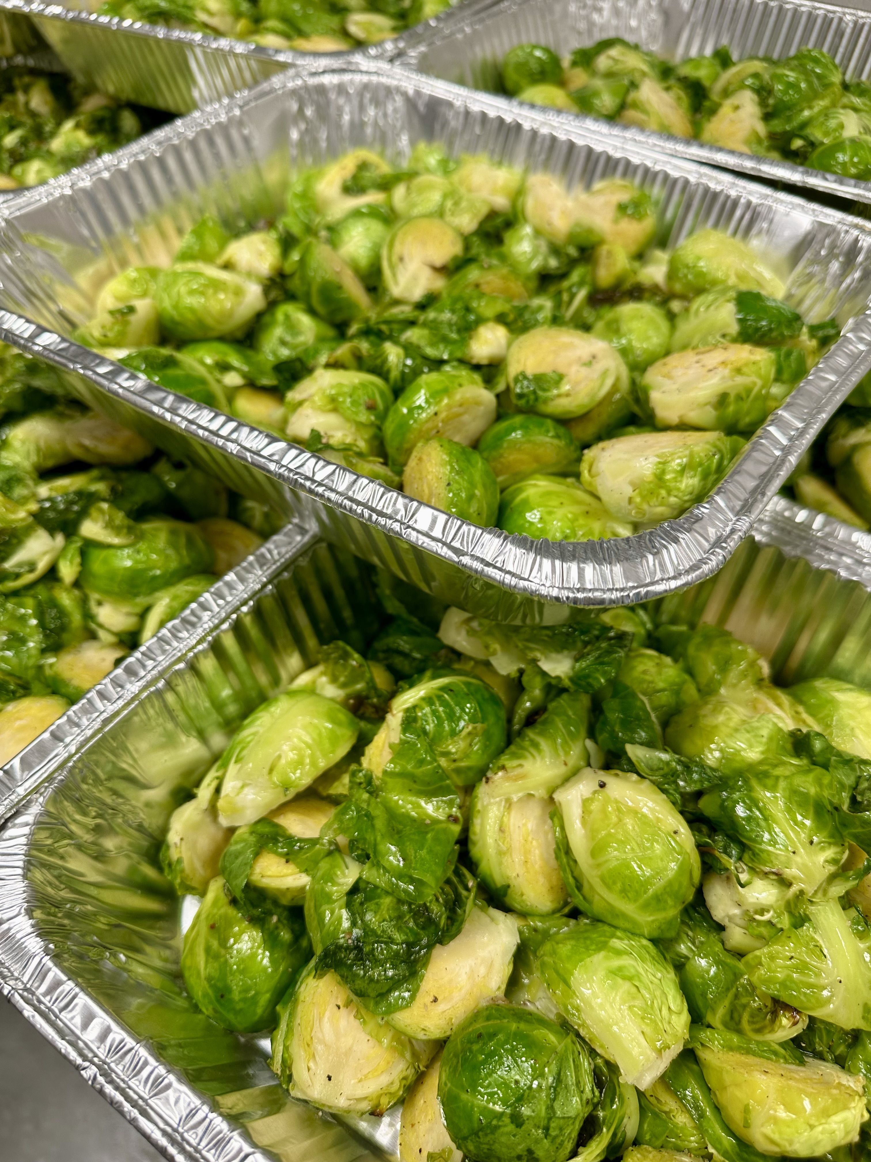 Close-up of aluminum trays filled with cooked, bright green Brussels sprouts seasoned with herbs and spices.