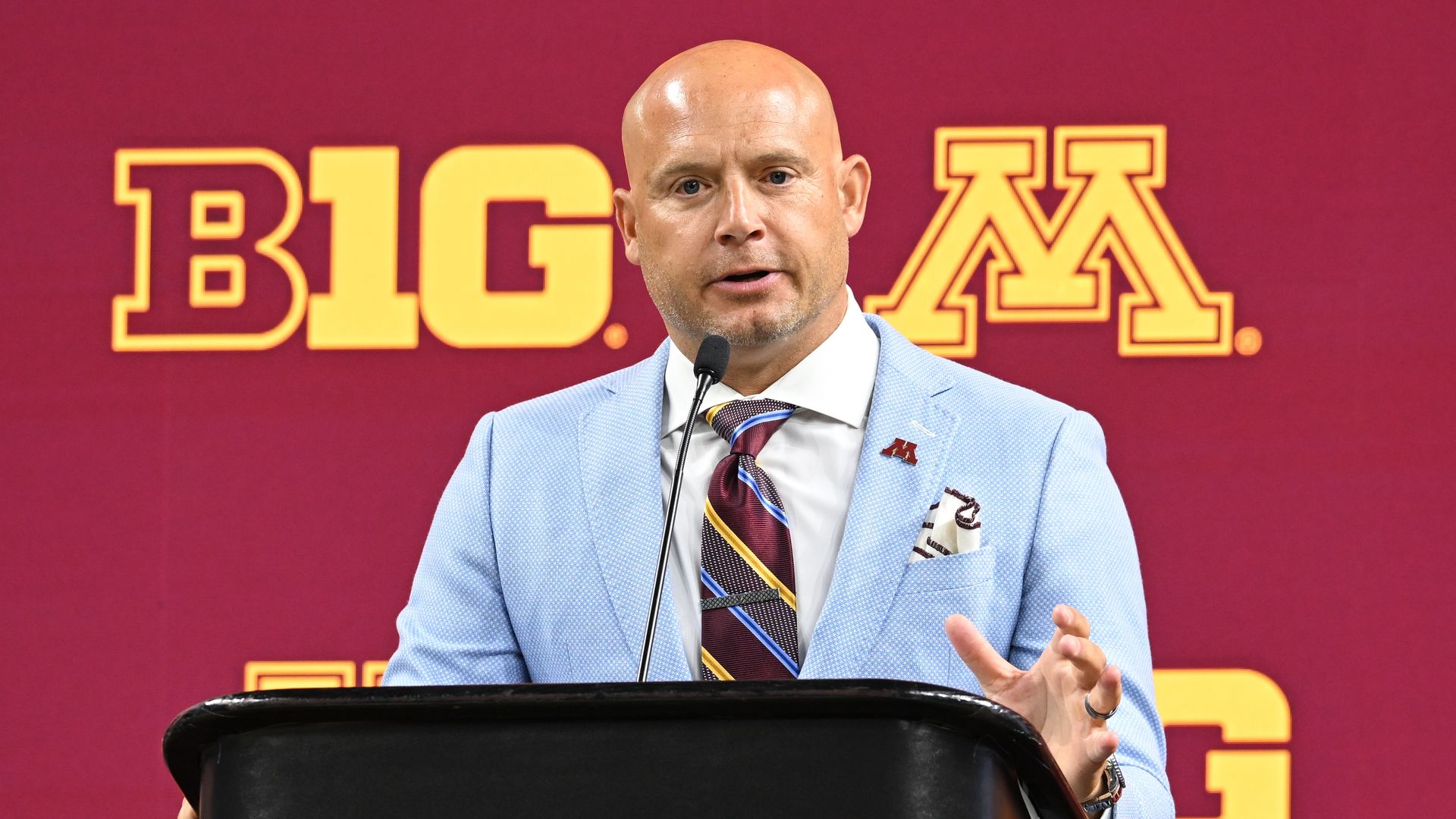 A bald man with light skin in a blue suit and a maroon striped tie speaks at a lectern with a backdrop featuring the logos of the Big Ten conference and the University of Minnesota