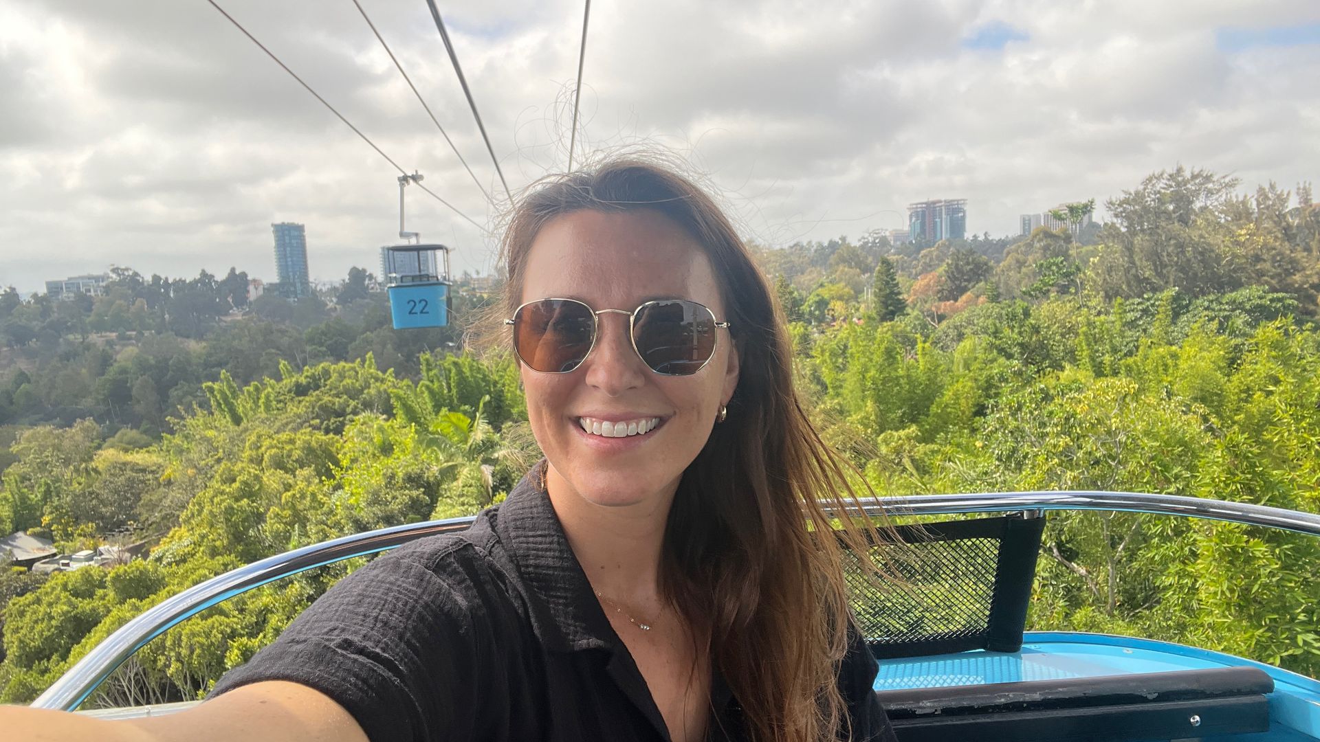 Kate, who has long brown hair and sunglasses ,smiling, taking a selfie on a blue cable car above lush green trees under a cloudy sky, with buildings in the distant background.