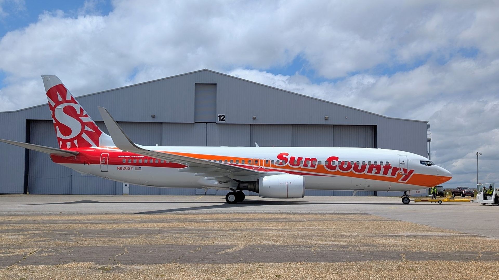 Side view of a Sun Country Boeing 737 parked on a tarmac in front of a large gray hangar labeled "12"; orange stripe along the fuselage, red tail design, engine under wing, ground crew nearby.