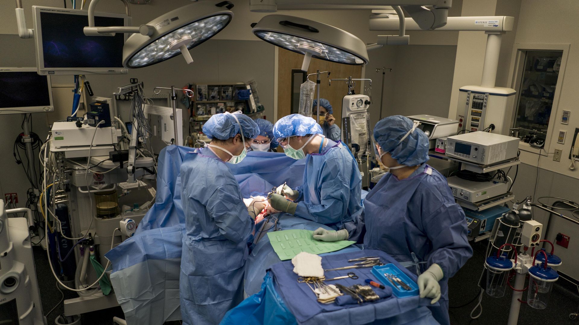 Surgeons and technicians in blue protective gear stand around a patient on an operating room table.
