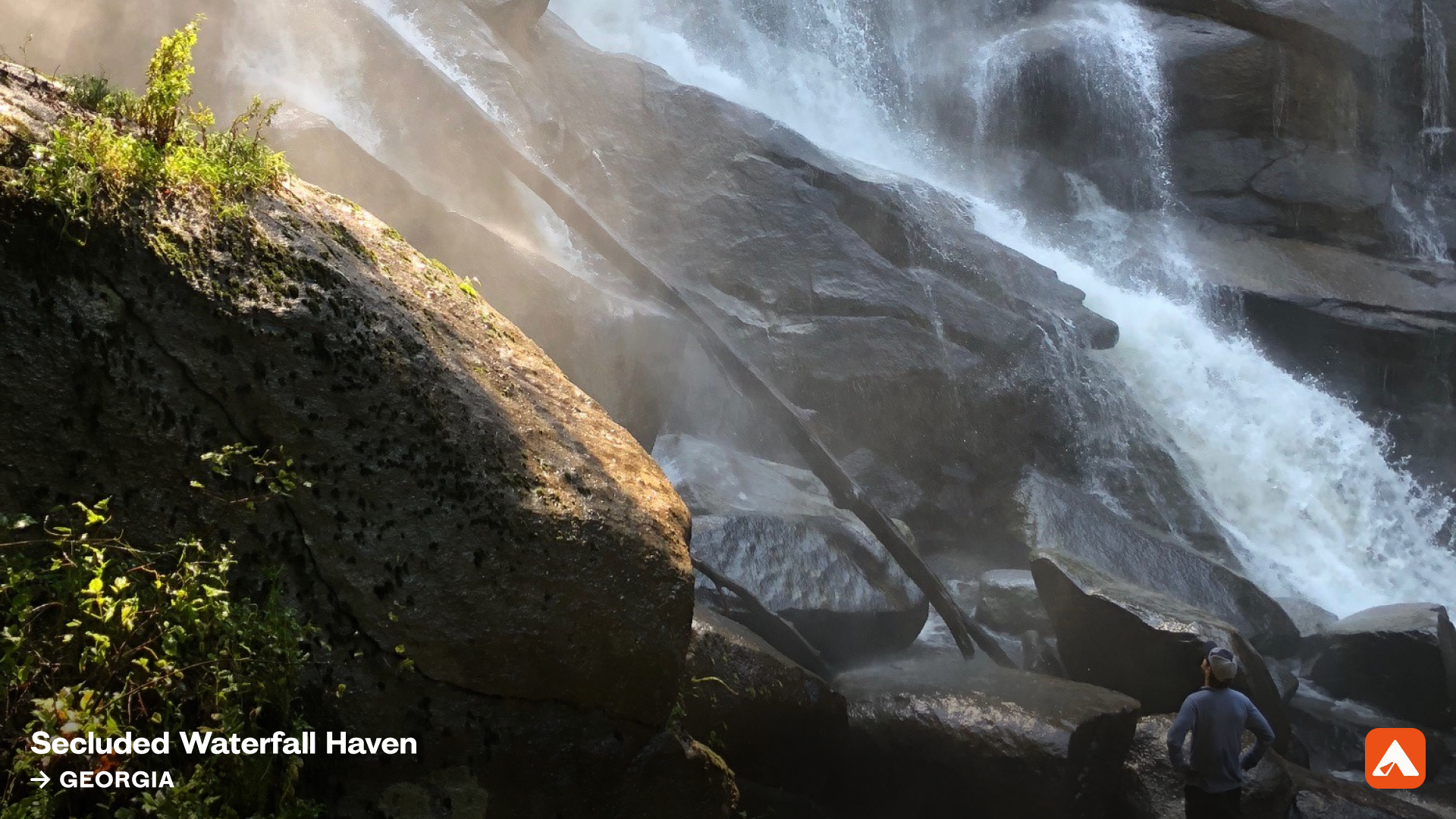 Person in blue shirt and cap stands near large mossy rocks at the base of a powerful waterfall surrounded by mist and greenery in Georgia's Secluded Waterfall Haven.