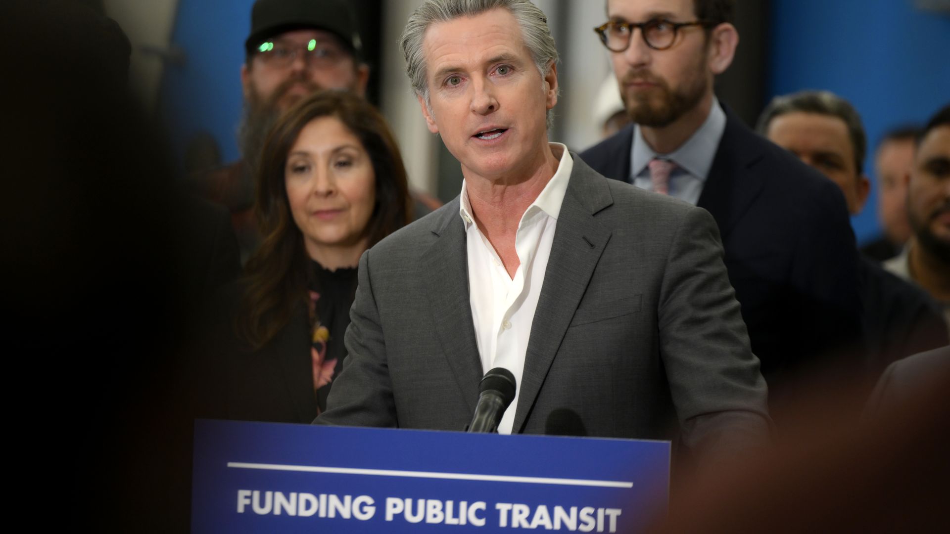 Man in gray suit and white shirt speaking at a podium with a sign reading "FUNDING PUBLIC TRANSIT," with several people standing behind him in a blurred background.