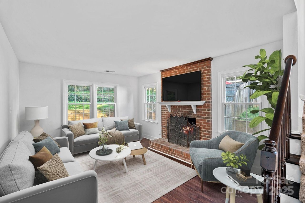 Cozy living room with two light gray sofas, a gray armchair, a brick fireplace with a TV above, white walls, large windows, indoor plants, and a round white coffee table on a light rug.