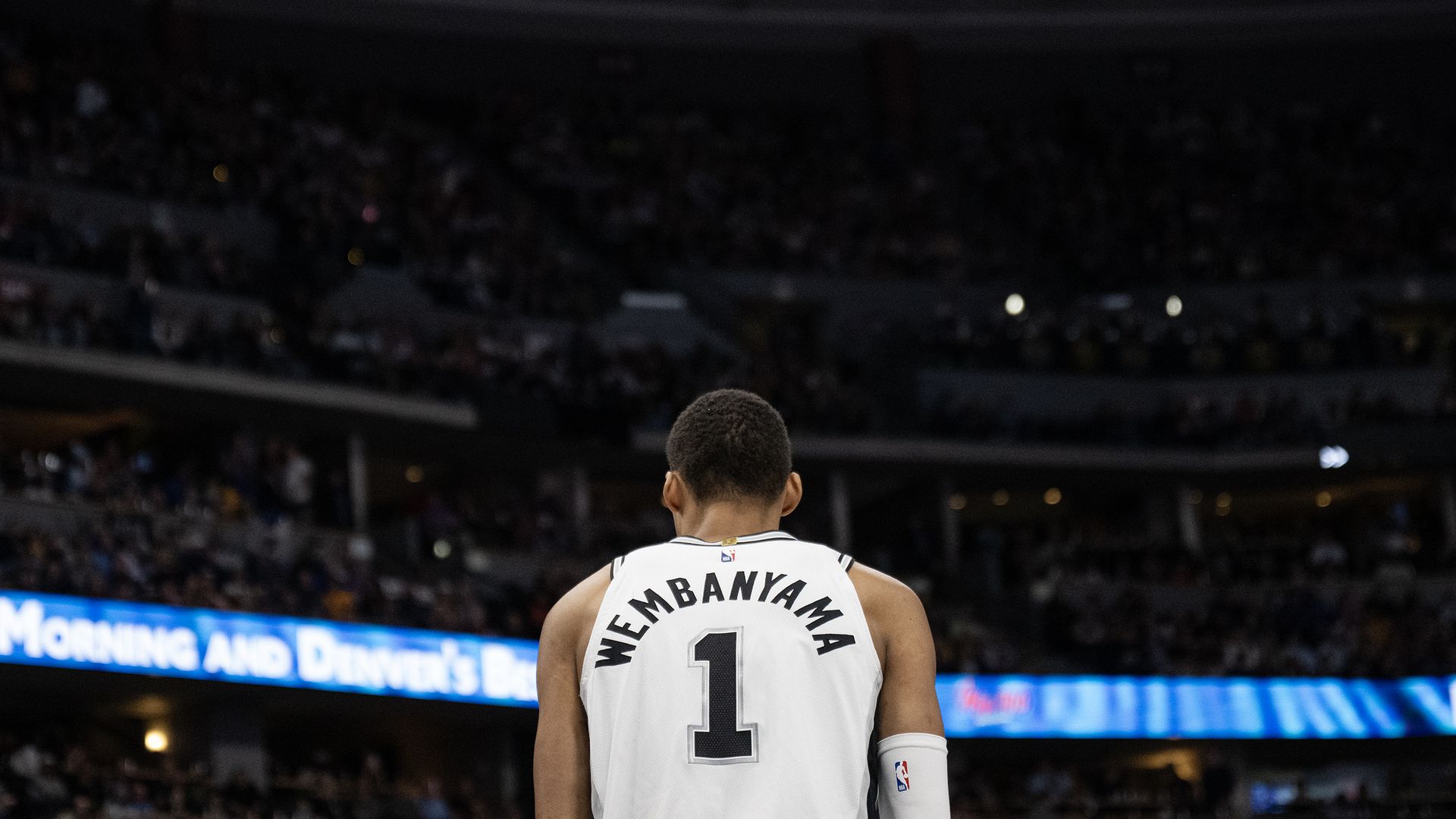 Back view of a basketball player in a white jersey with the number 1, standing on a court in a packed arena; a bright blue LED banner runs along the stands behind him.