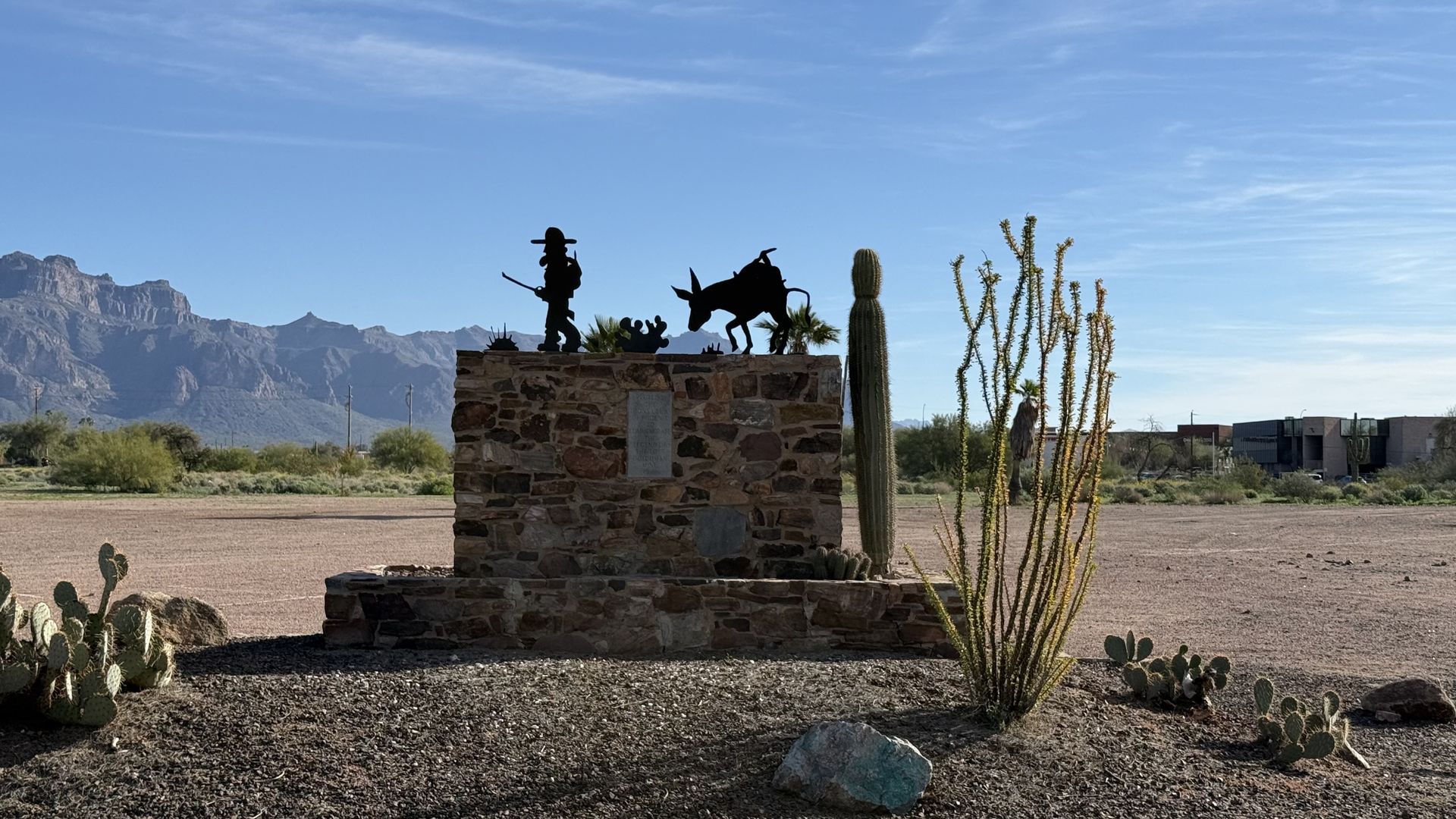 Metal silhouette statue of a man and donkey both walking on a stone pedestal in a desert landscape with mountains in the background.
