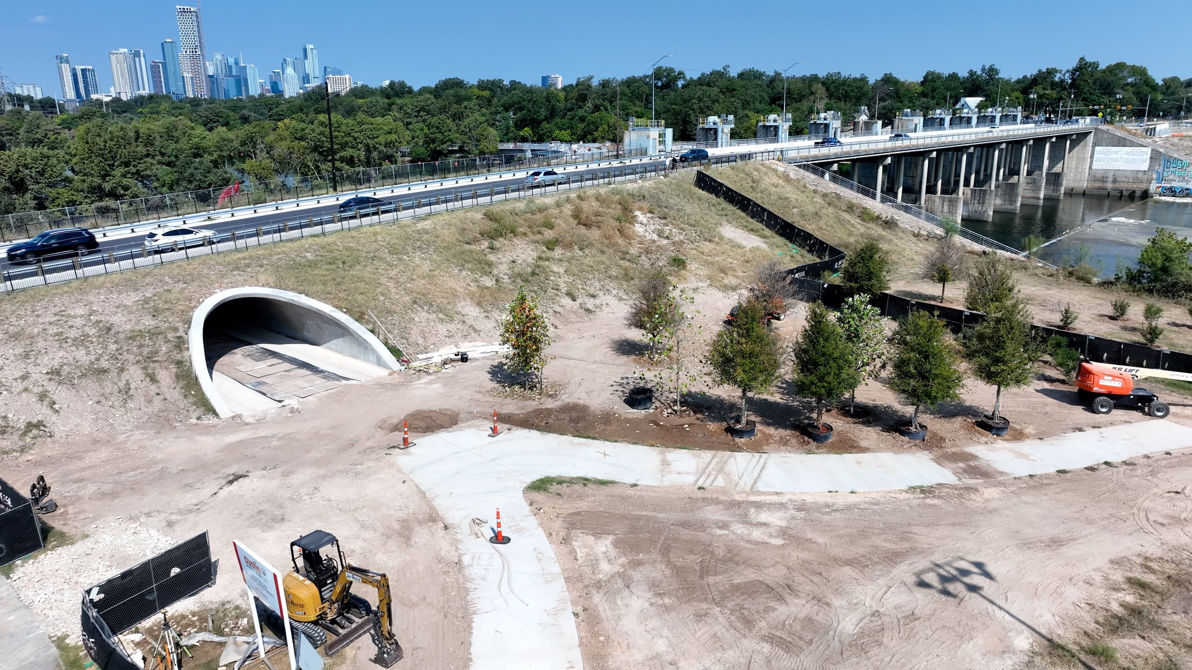Construction site with excavator, newly planted trees in pots, concrete path, tunnel entrance, bridge over river, and city skyline in the background under a clear blue sky.