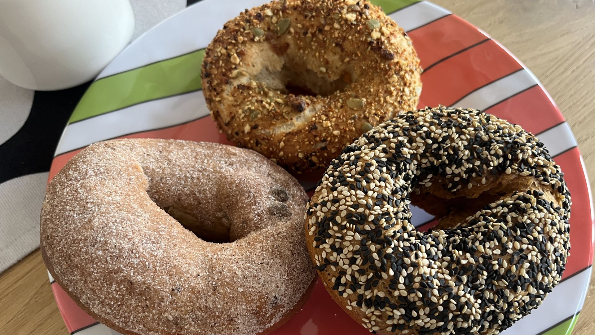 Three bagels on a colorful plate: one coated with mixed seeds, one with cinnamon sugar, and one with assorted nuts and seeds, with part of a white mug visible on the side.