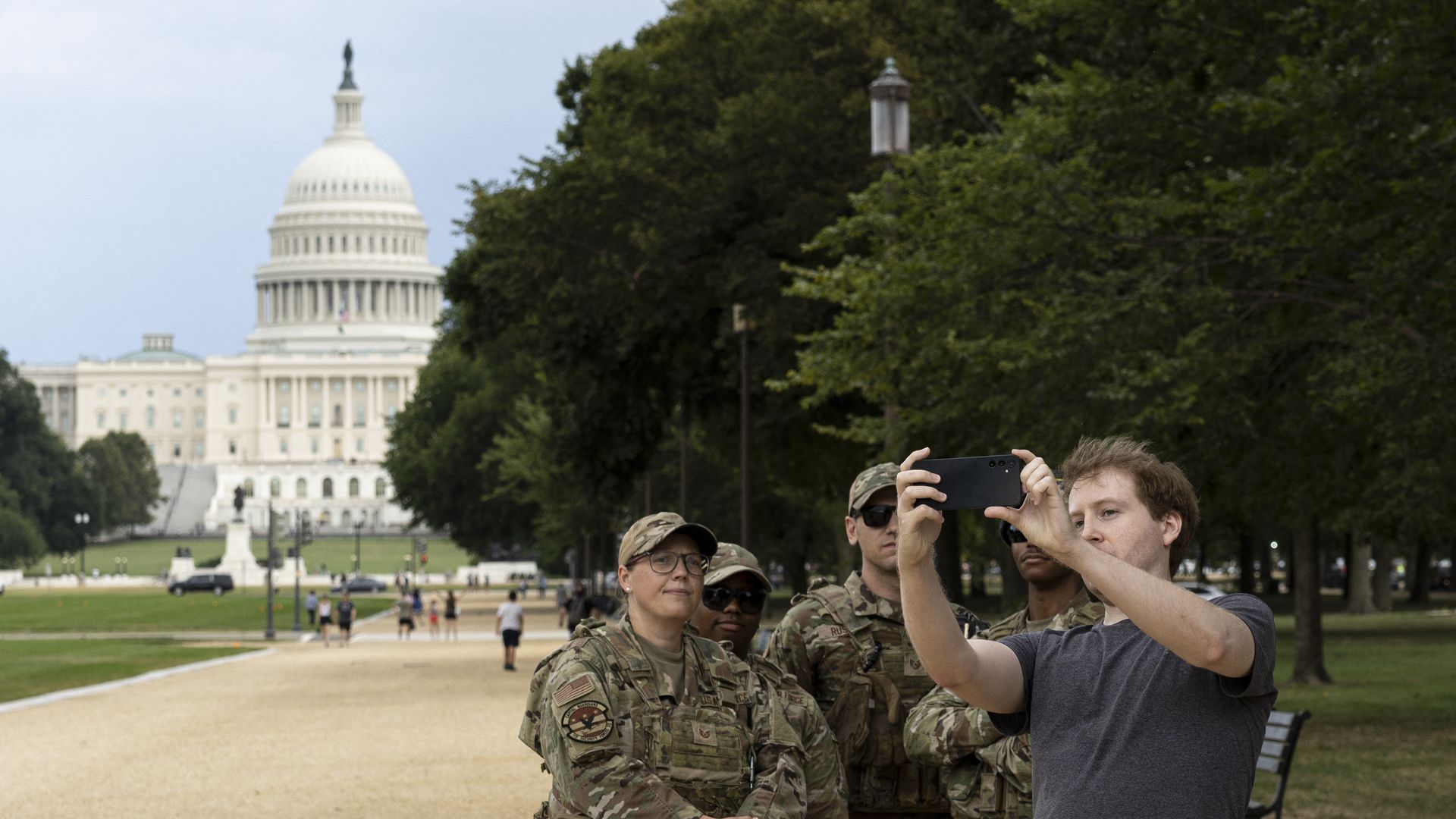 A man in a gray shirt takes a selfie with four military personnel in camouflage uniforms near the U.S. Capitol building on a gravel path lined with trees and benches.
