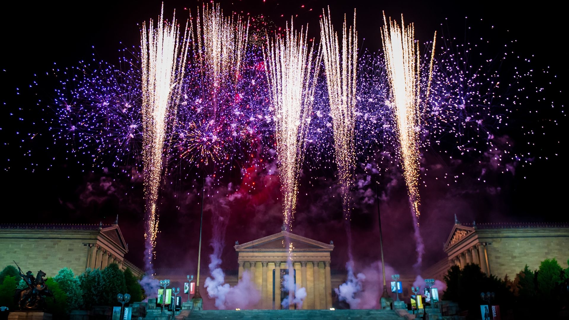 Yellow, red and purple fireworks going off at night in front of a large museum styled after a Greek temple. 