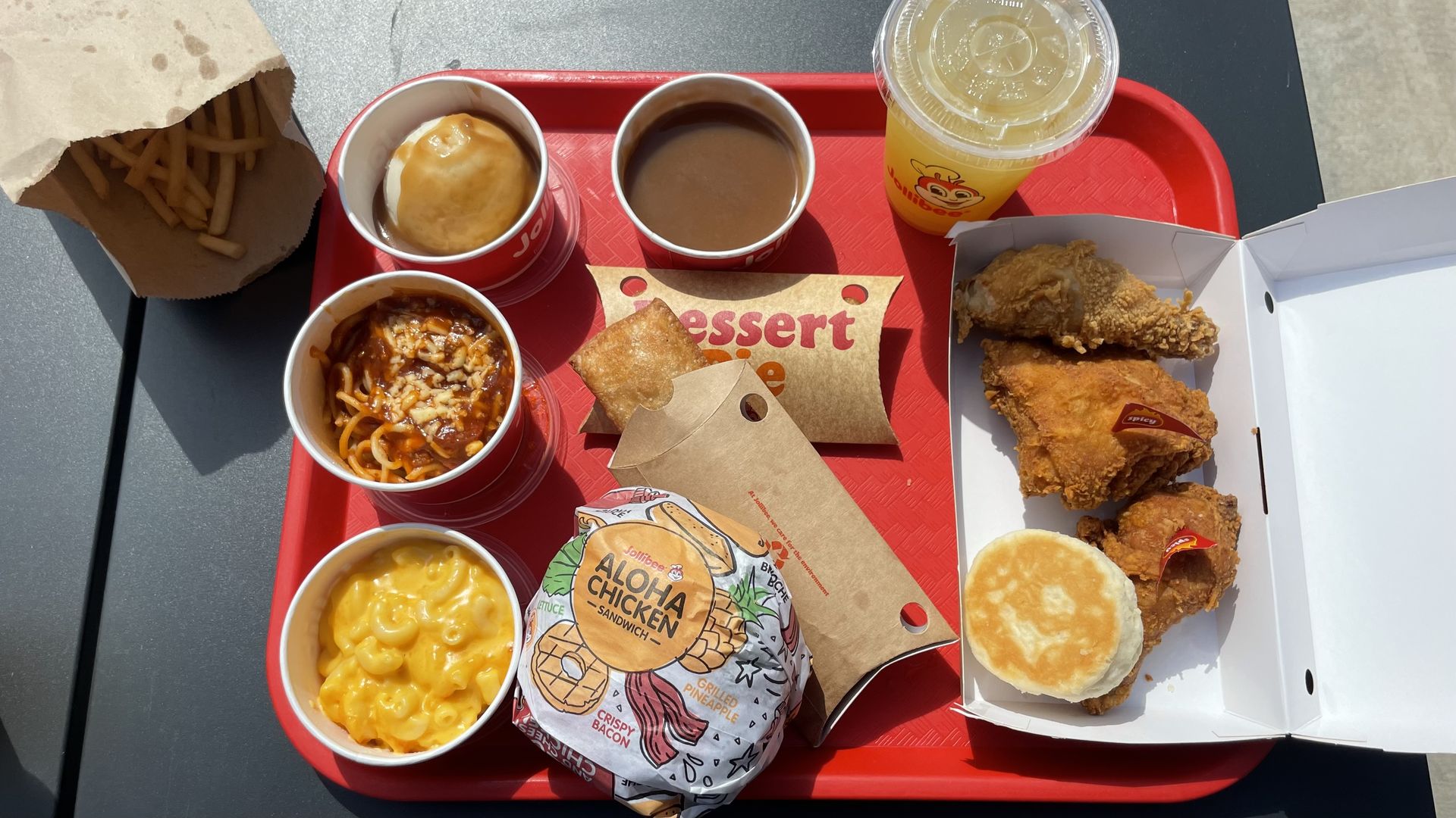 Red tray with fast food including fries, mashed potatoes with gravy, spaghetti, mac and cheese, Aloha chicken sandwich, dessert pie, fried chicken pieces with spicy labels, biscuit, and yellow drink.