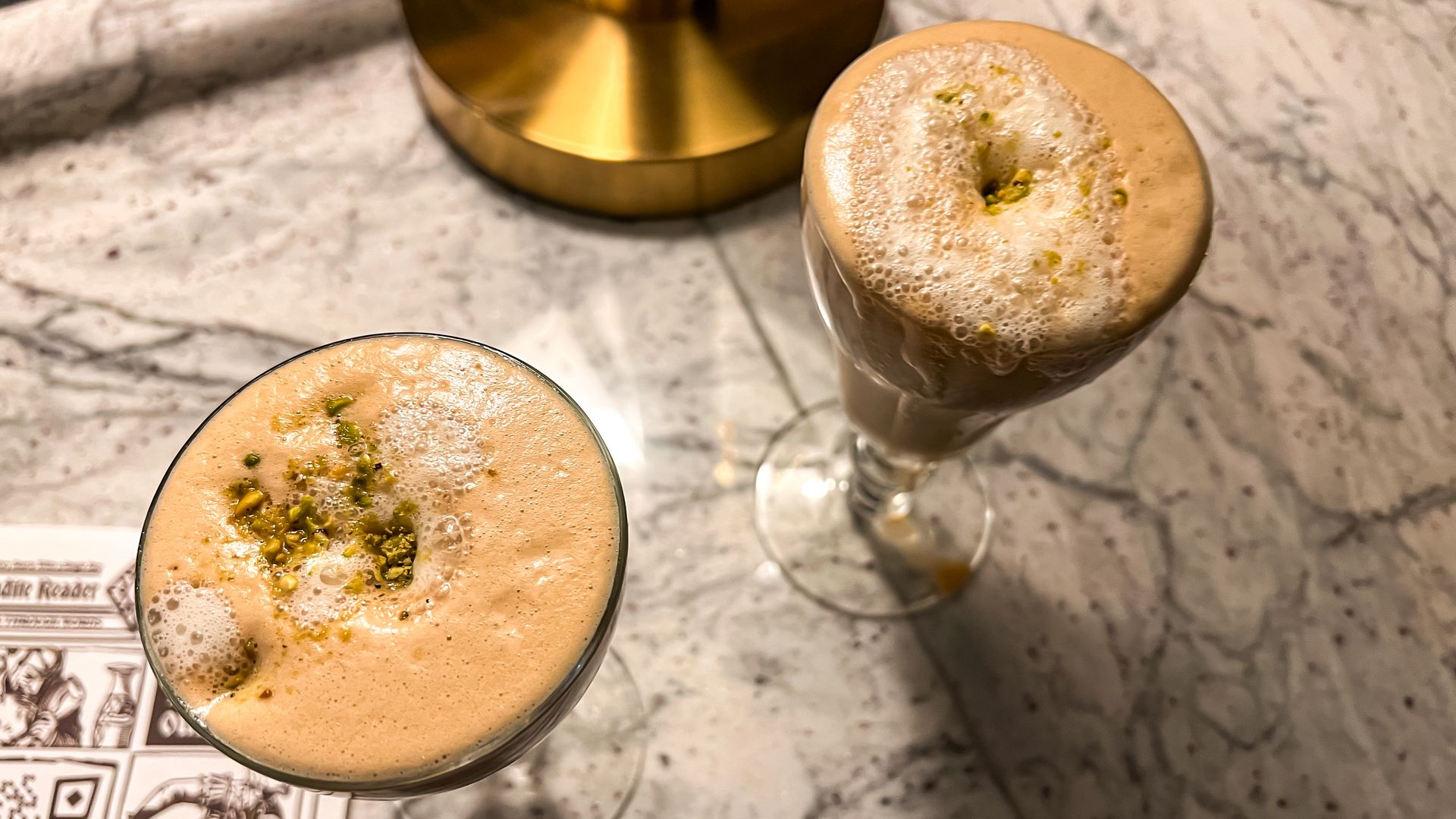 Two cocktails with foamy tops sit on a marble bar top. 