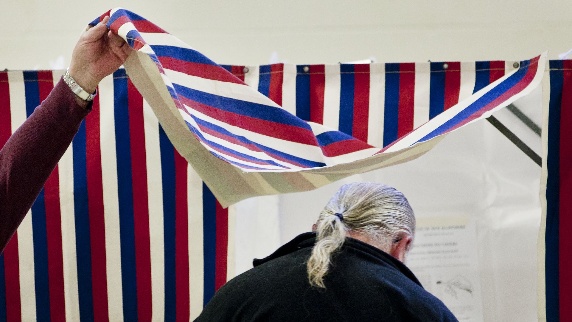 Someone lifts the red, white, and blue curtain as a man enters a polling booth