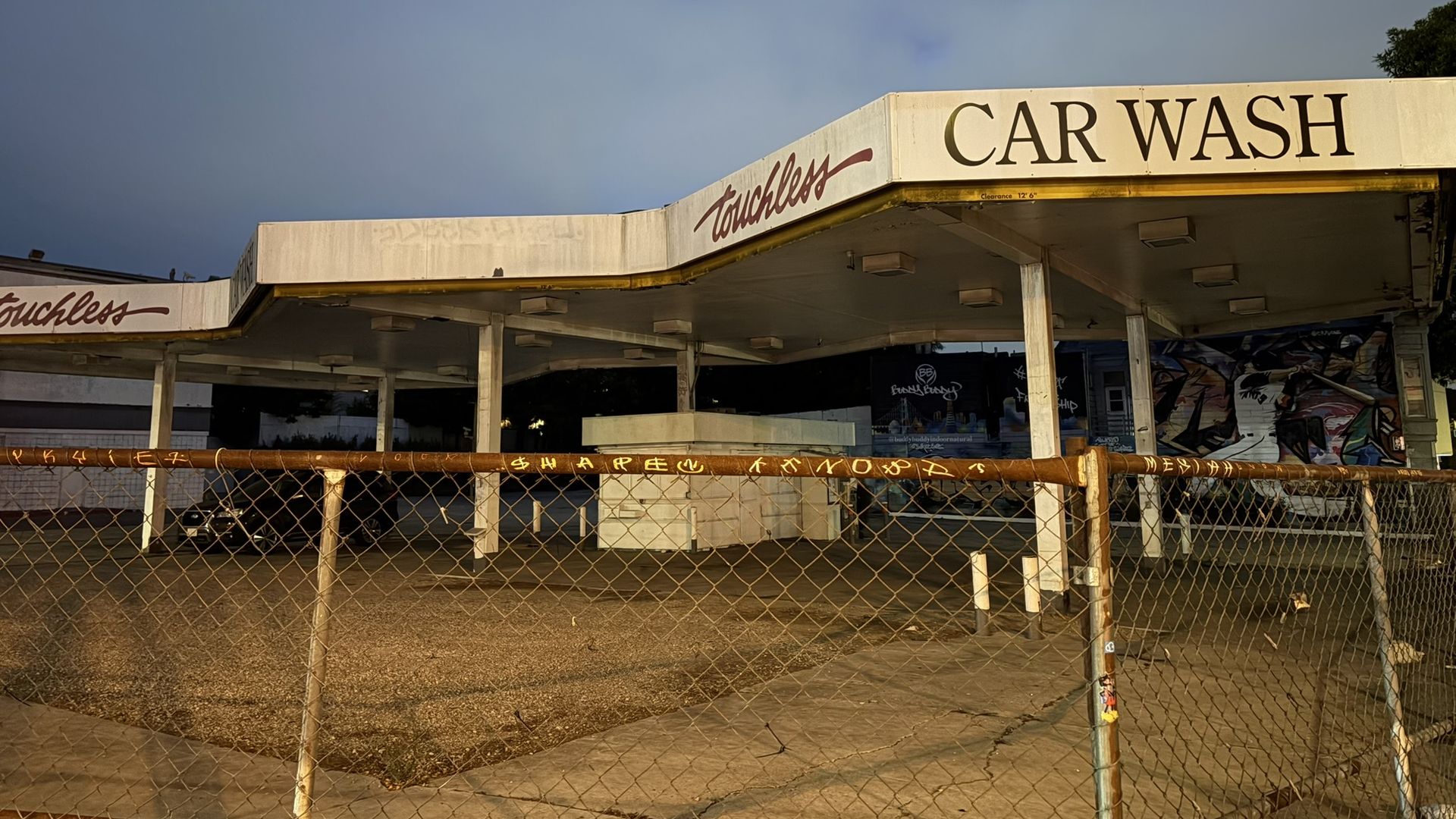Wide shot of a car wash with a large "CAR WASH" sign on a white canopy, chain-link fence in the foreground, graffiti on the far wall, and a blue sky with a tree to the right.