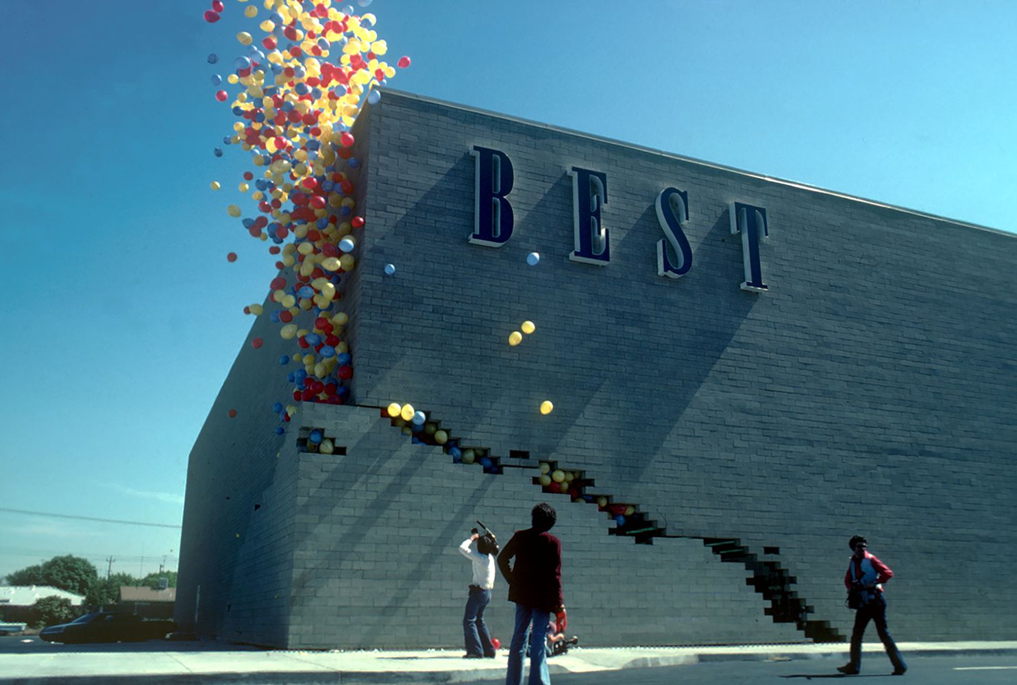 Blue brick building with a large BEST sign. Multicolored balloons spill from the top left, cascading down the wall along a stepped opening, while three people stand on the sidewalk below.