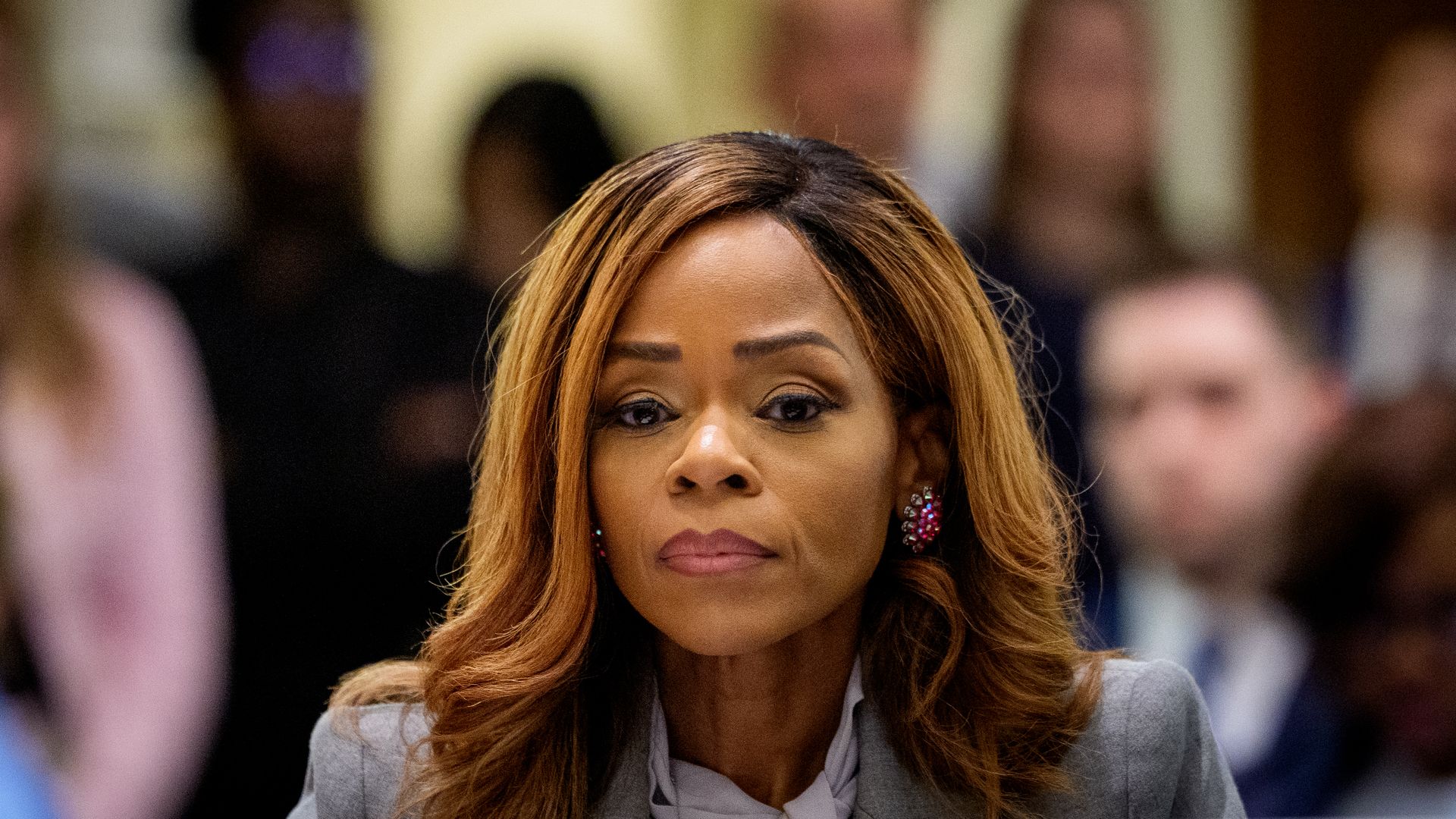 Close-up of a woman with light brown hair and pink lipstick, wearing a gray blazer and pink earrings, surrounded by a blurred crowd in a warmly lit indoor setting.
