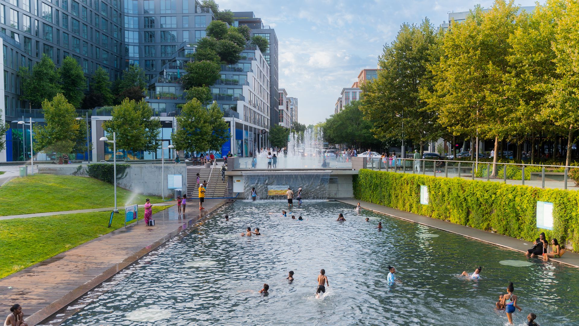 Urban waterfront park with a long shallow pool, people swimming and playing in clear water, modern glass apartment buildings line the left, lush trees and a walkway on the right, a central fountain.