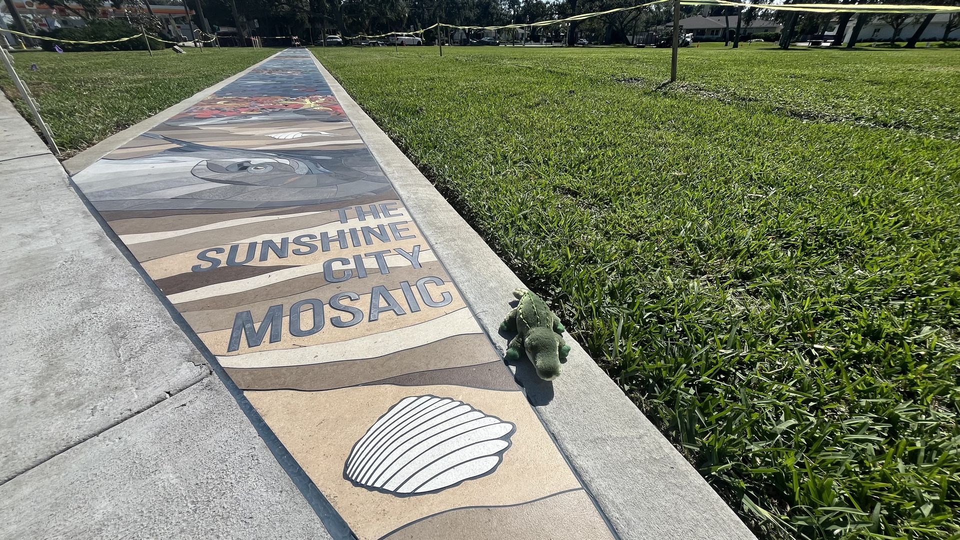 Mosaic sidewalk with text "The Sunshine City Mosaic" and a shell design, bordered by grass on the right where a small green toy alligator lies in the sun, with trees and houses in background.