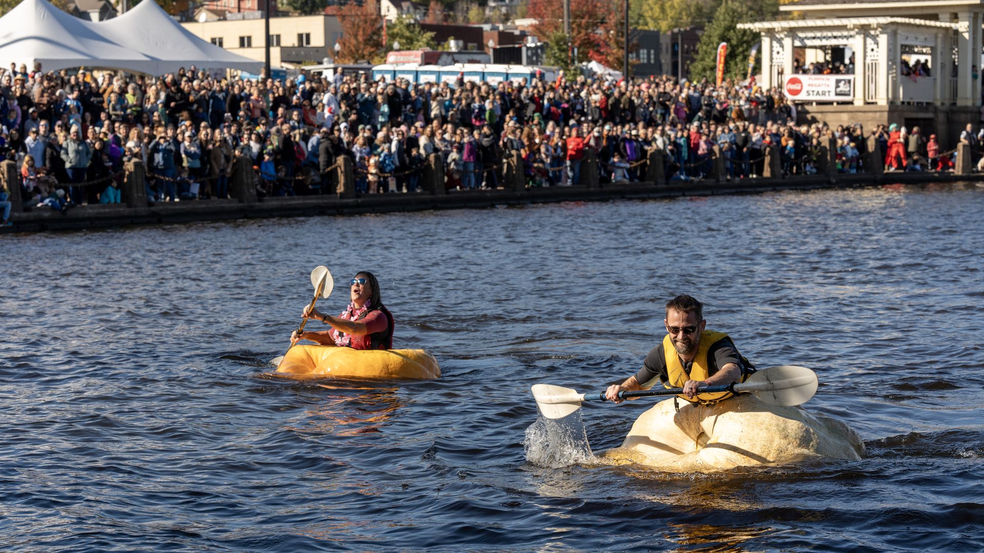Two people paddling in boats shaped like pumpkins on a river, with a large crowd watching along the shore on a sunny day with fall foliage.