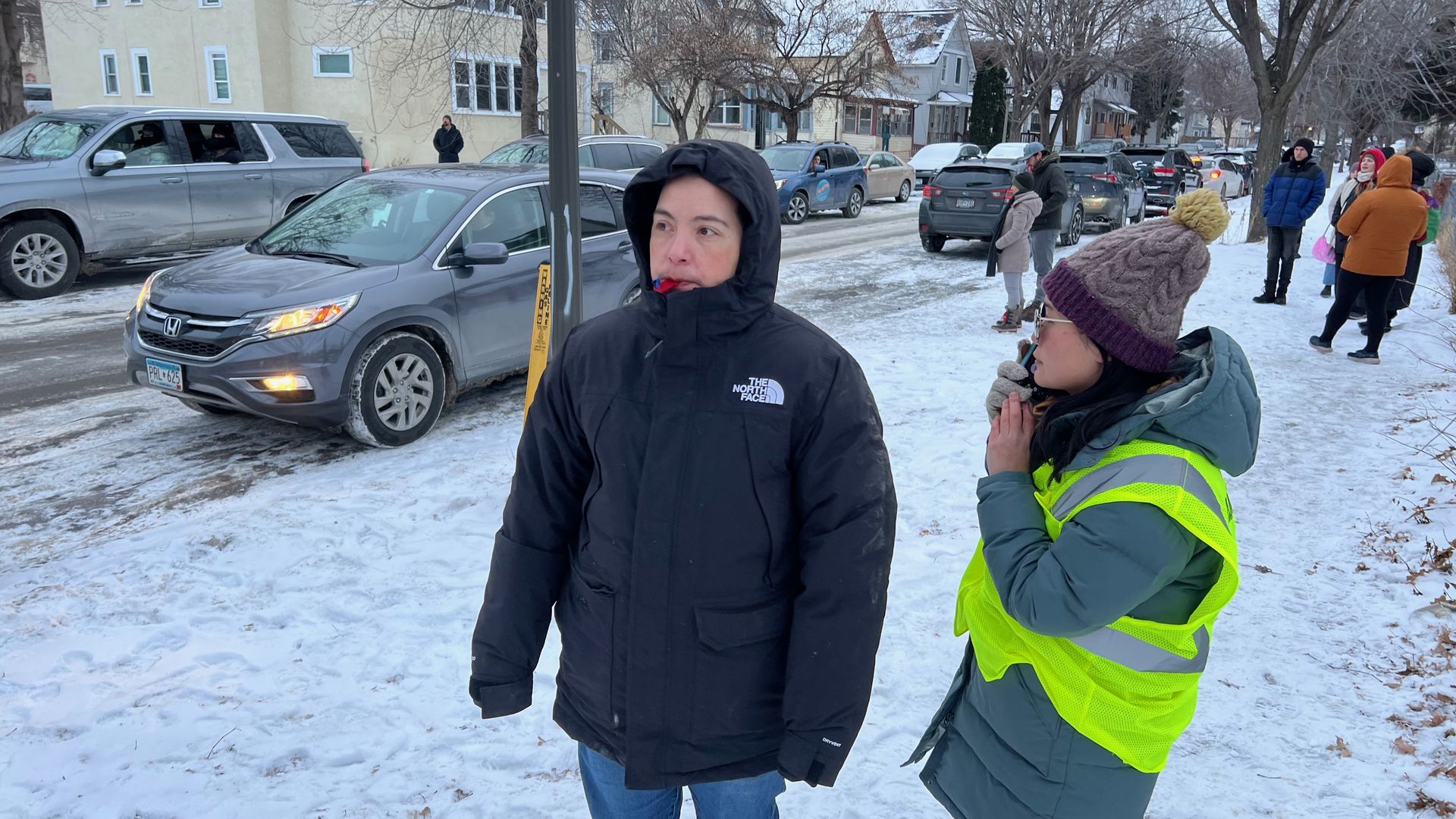 Two people bundled in winter clothes stand on a snowy street; one in a black North Face jacket, the other in a green coat with a yellow safety vest and knit hat, cars and people line the road.