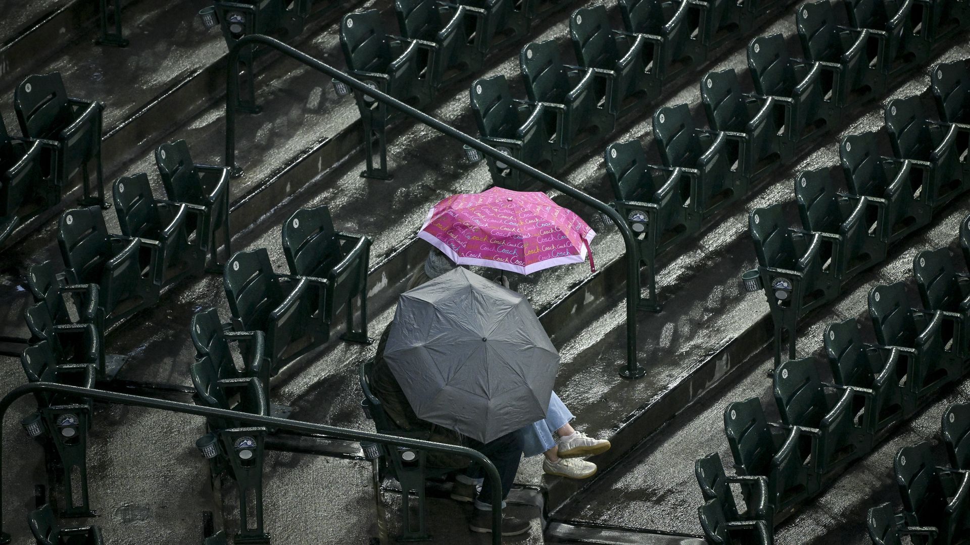 Rainy stadium seating with many green rows; a pink patterned umbrella and a gray umbrella sit on the wet steps, with a person's legs and sneakers visible beneath the pink shade.