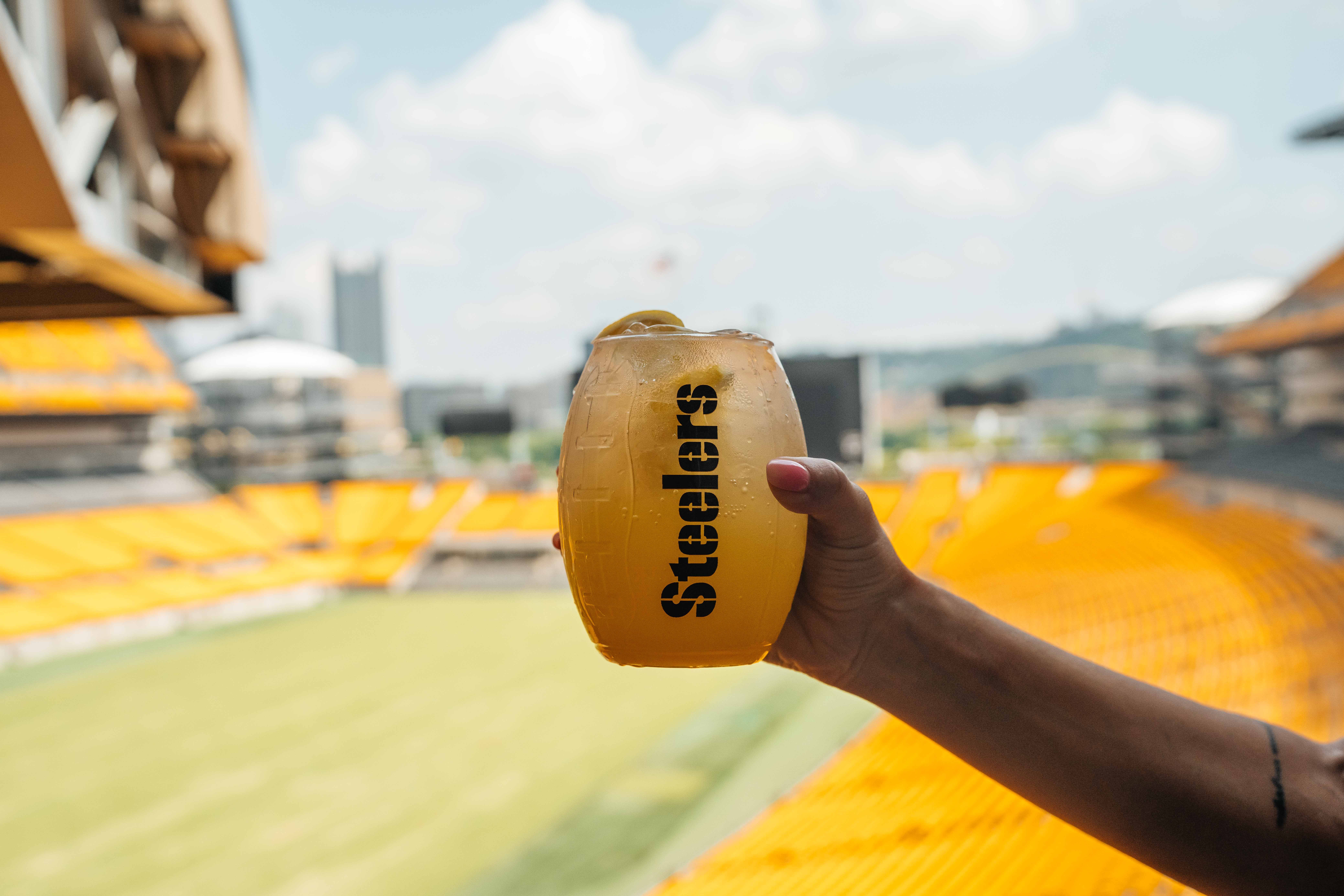 A hand holds an orange beverage in a clear cup with ice, labeled "Steelers" in the foreground. Yellow stadium seating and a bright blue sky with clouds blur in the background.