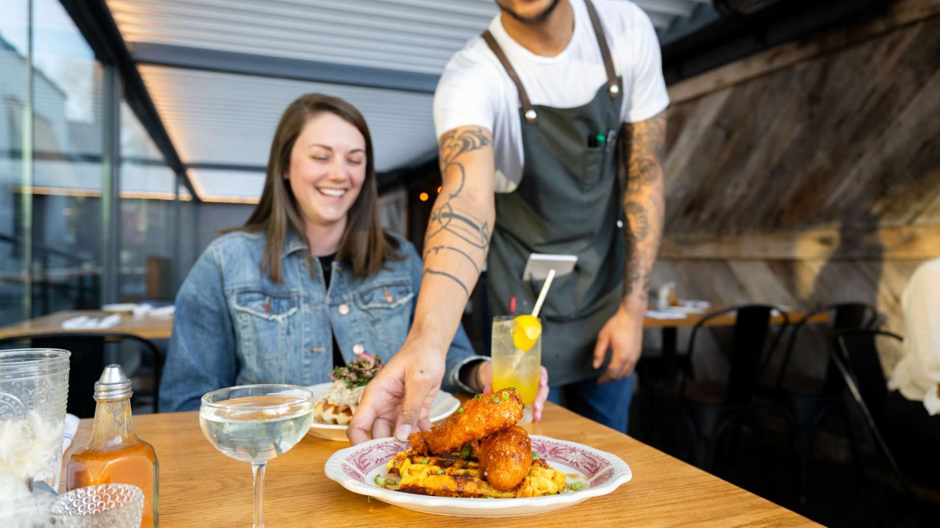 Waiter with tattoos serves a plate of fried chicken and waffles to a smiling woman in a denim jacket at a wooden table in a casual restaurant.