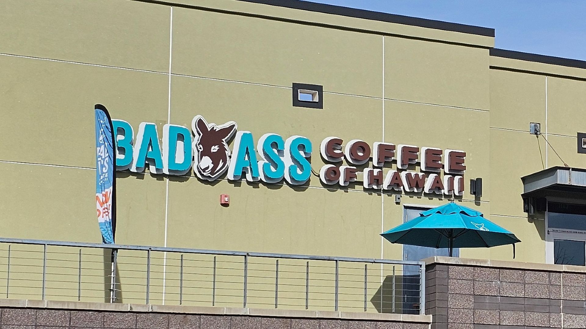 Exterior of a building with a sign reading "Bad Ass Coffee of Hawaii" in blue and brown letters, featuring a donkey logo and a blue patio umbrella nearby.