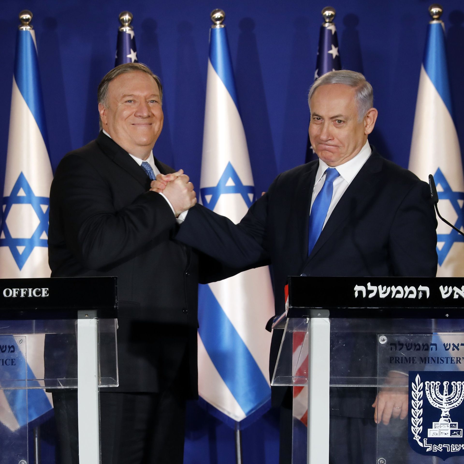 Mike Pompeo and the Israeli prime minister shake hands in front of a row of Israel and American flags, standing behind two podiums. 