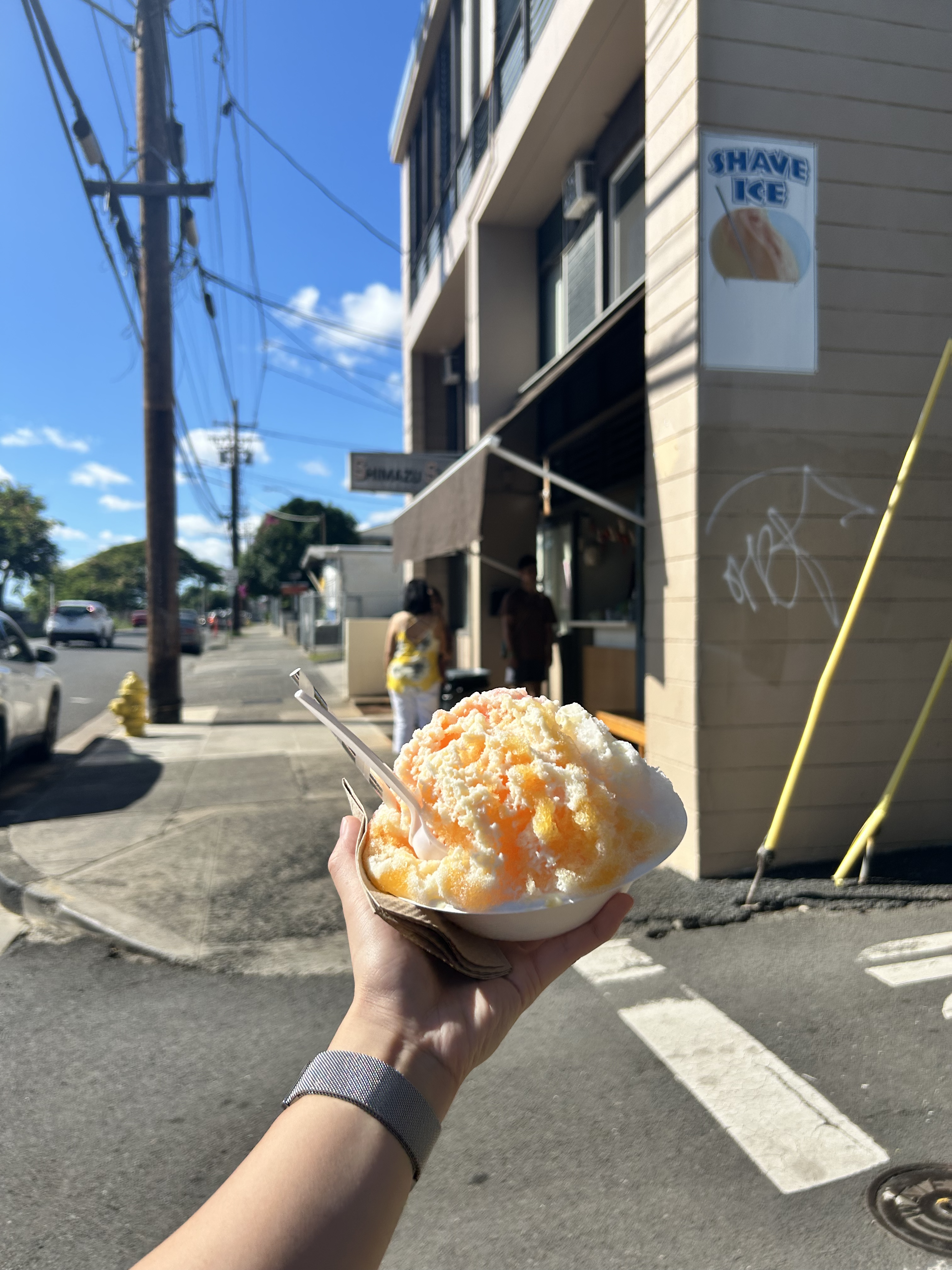 Photo of a hand holding a golden and white shaved ice bowl in front of a roadside shop