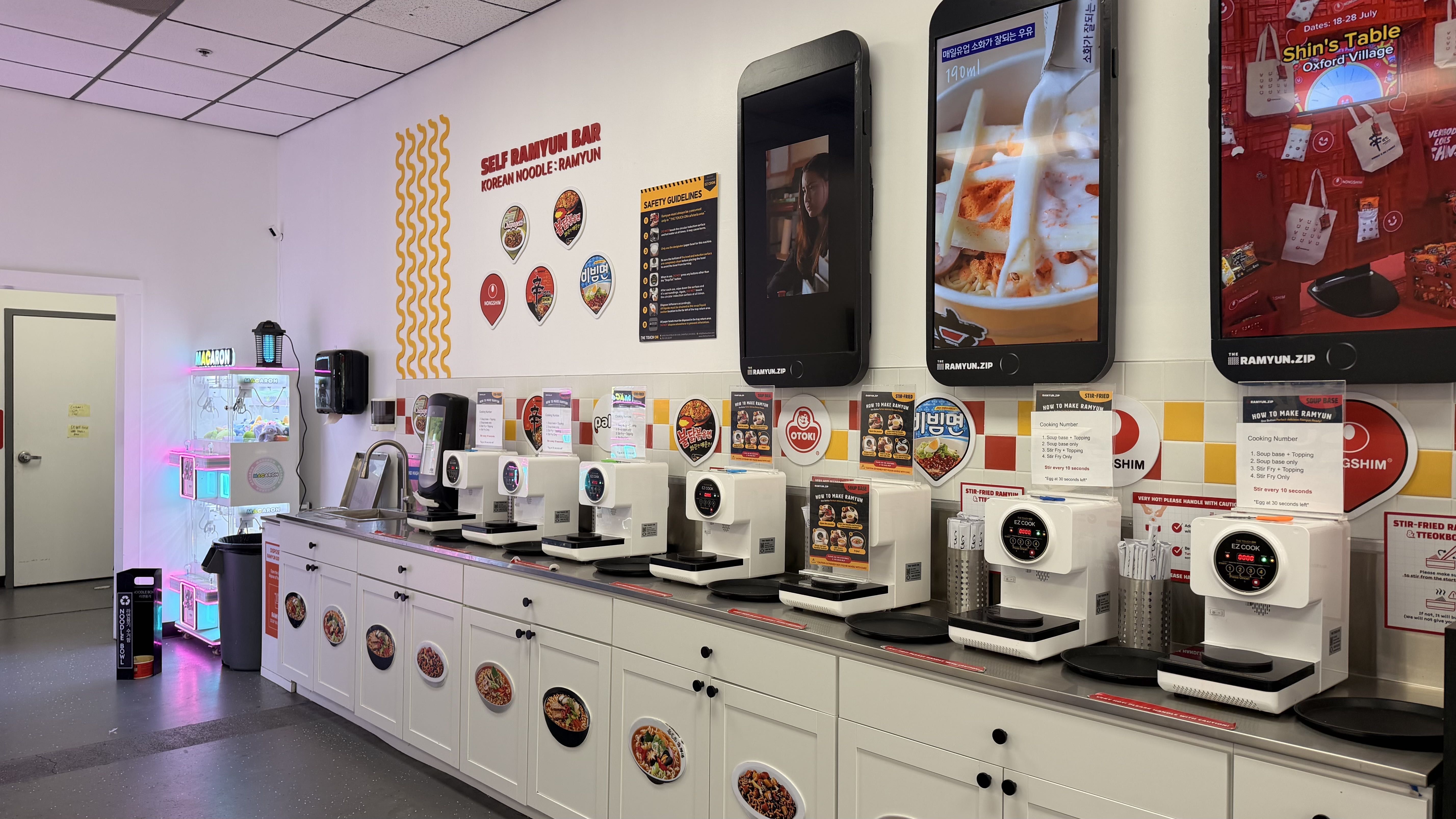 Bright self-serve ramen shop with a white counter and appliances, three large wall screens showing ramen images, and a "Self Ramyun Bar" sign with colorful decals along the wall.
