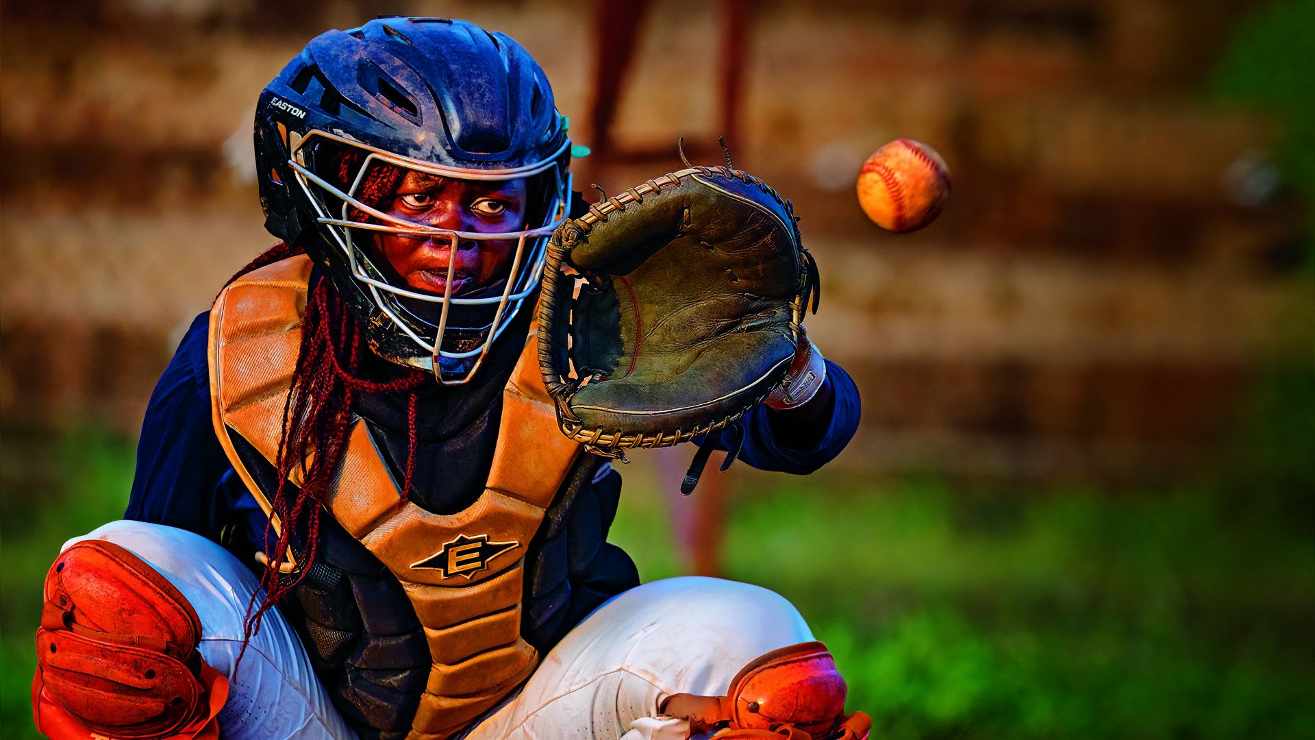 Baseball catcher in a blue helmet and orange protective gear extends a gloved hand to catch a speeding ball, eyes visible through the mask against a blurred field.