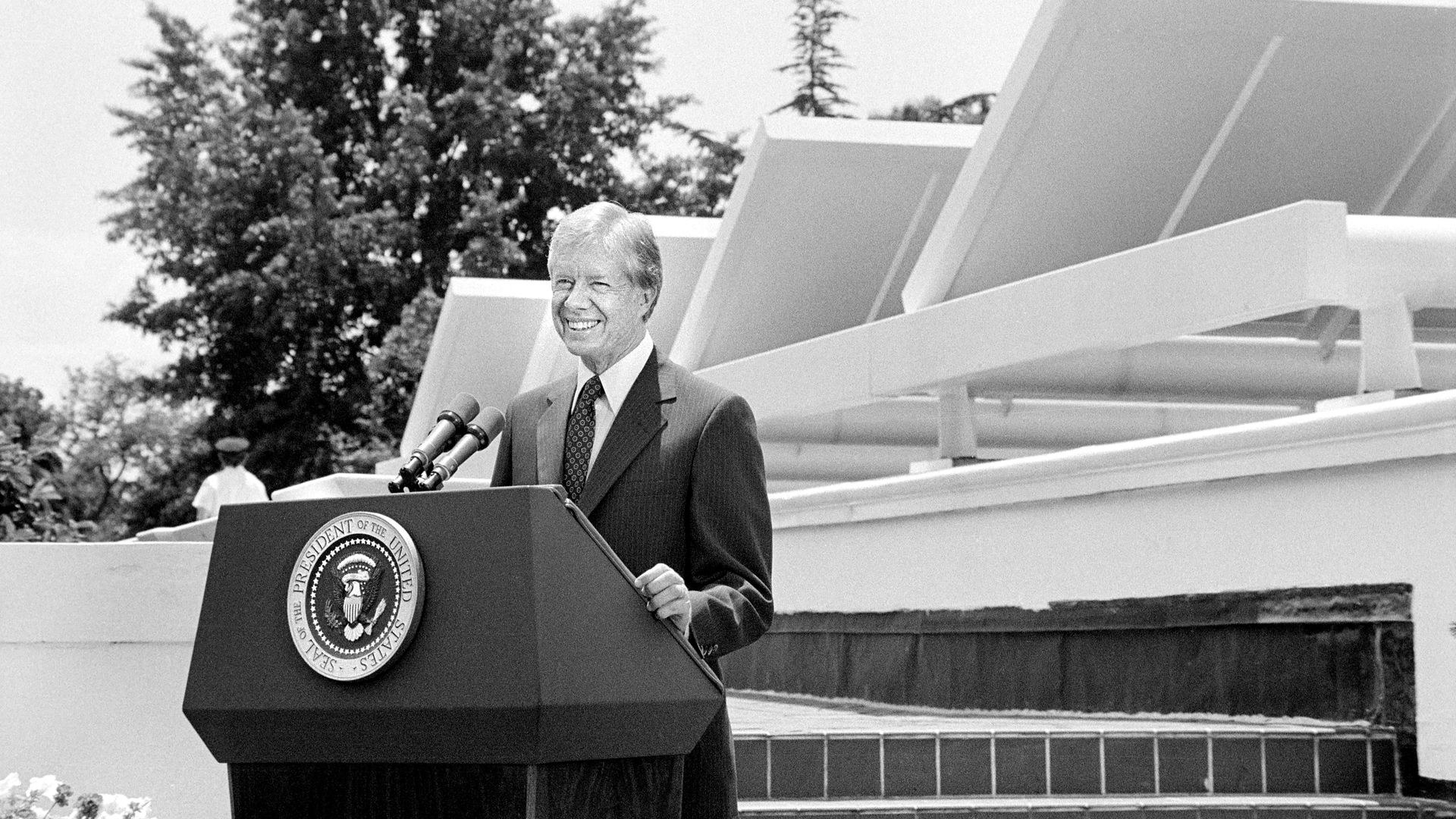 Jimmy Carter in a black and white photo from 1979, standing behind a lectern with the presidential seal, with multiple solar panels behind him.
