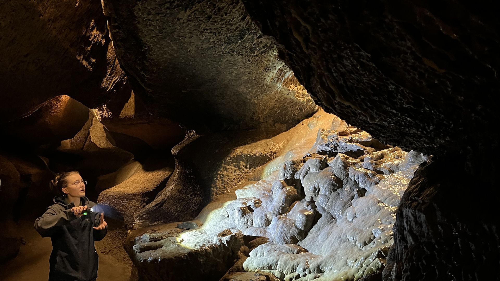 A woman with a flashlight illuminates a slick-looking rocky formation in a dark cave