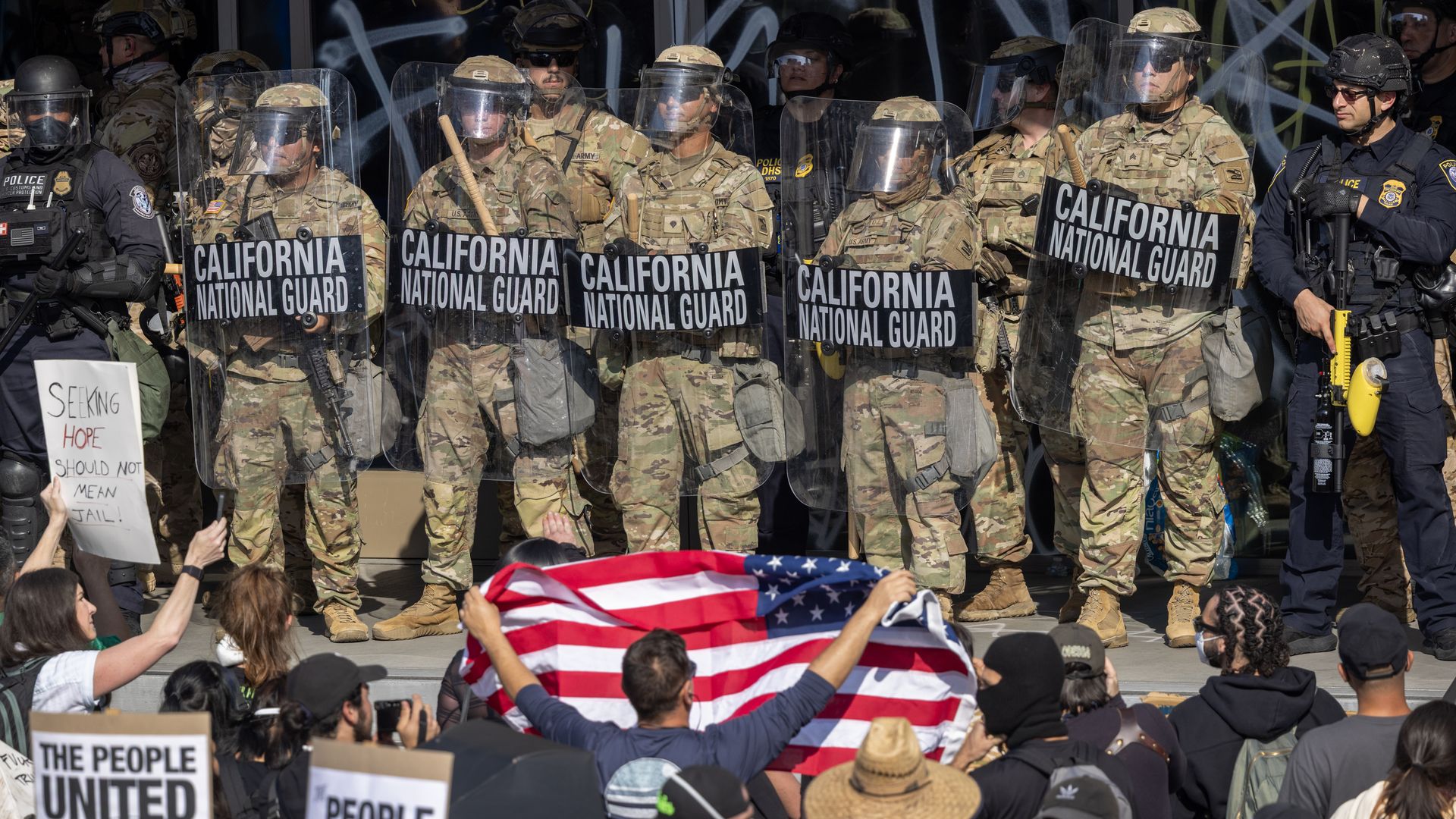 Protesters hold signs while one lifts an American flag while California National Guardsmen stand guard.