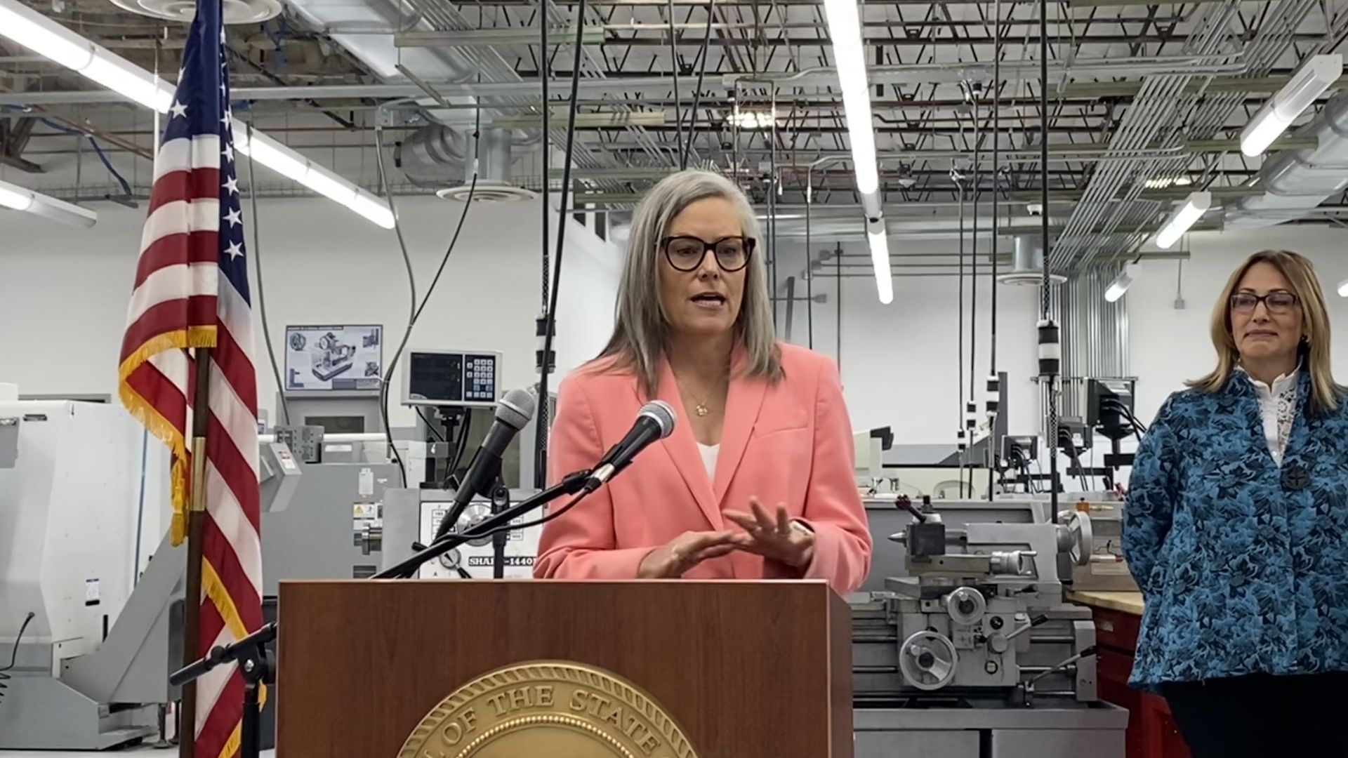 Katie Hobbs speaks at a lectern with industrial equipment in the background.