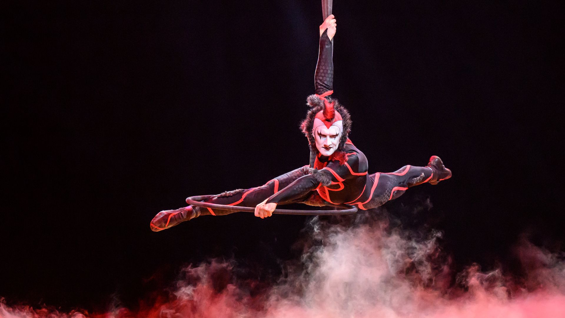 A Cirque du Soleil performer in a red and black costume hangs above the stage, which is covered in smoke.