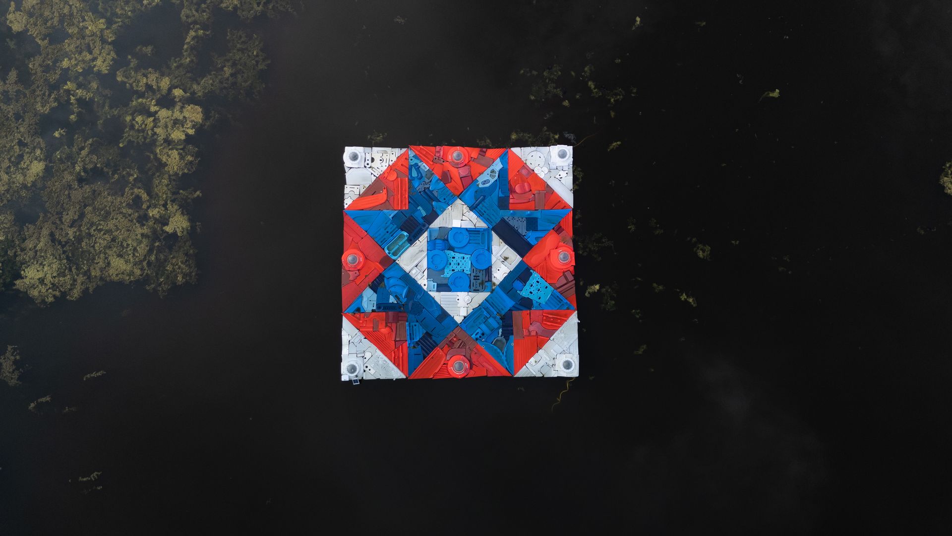 An aerial view of a square floating platform covered in red, blue, and white geometric patterns, surrounded by dark water and green tree-covered banks.